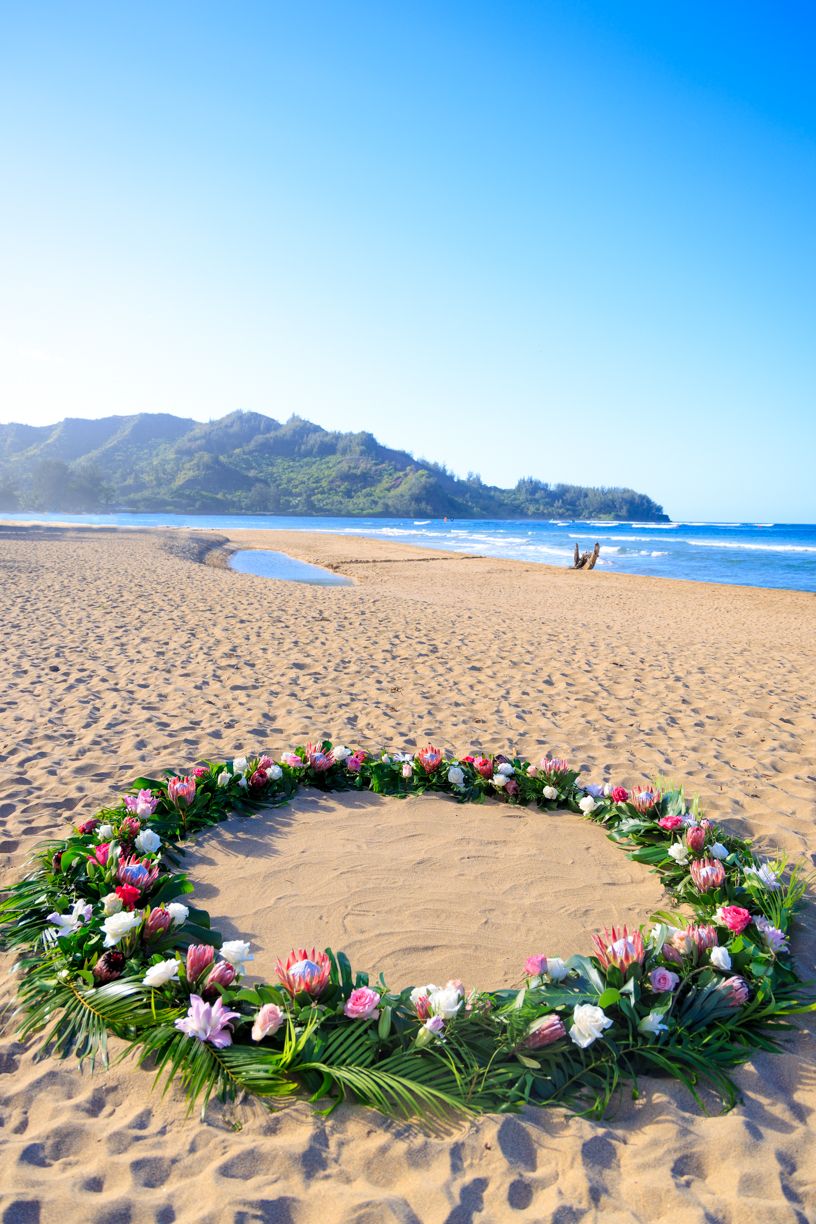A circular arrangement of tropical flowers and palm fronds on a sandy beach with mountains in the distance.