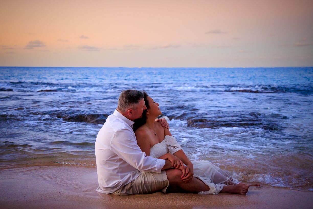Couple embracing on beach at sunset with ocean waves in background.