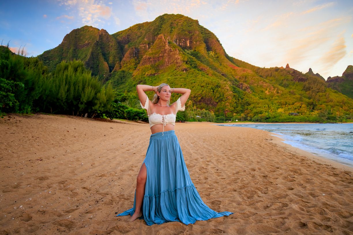 Flowing blue dress on sandy beach with towering green mountains and sunset sky.