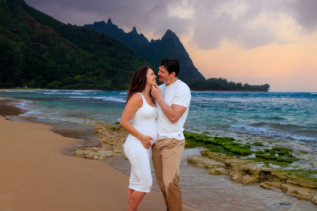 Two people share a romantic moment on a scenic beach with dramatic cliffs and sunset sky.