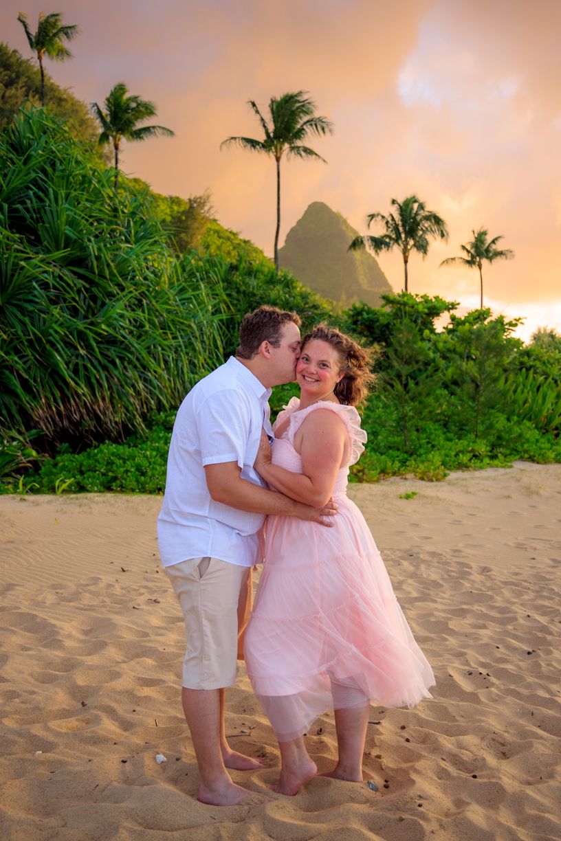 Couple sharing romantic moment on sandy beach with palm trees and mountains during golden hour.