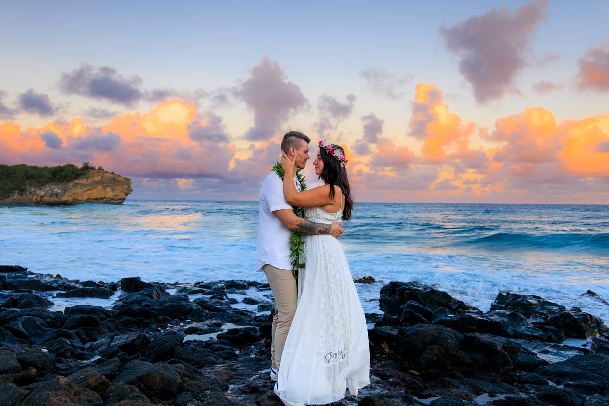 Wedding photos on rocky Hawaiian coastline during vibrant sunset with crashing waves.