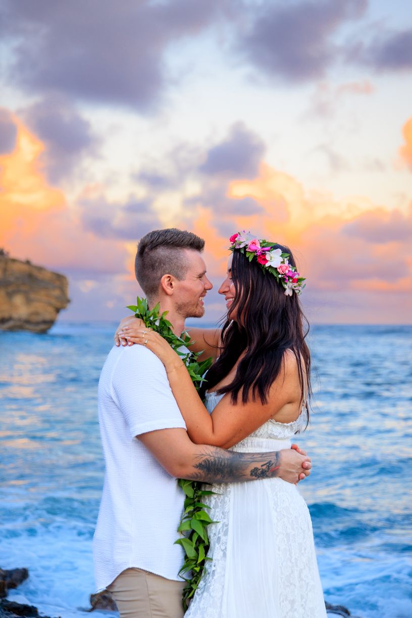 A romantic couple shares intimate moments during sunset at a Hawaiian beach with crashing waves and golden skies.