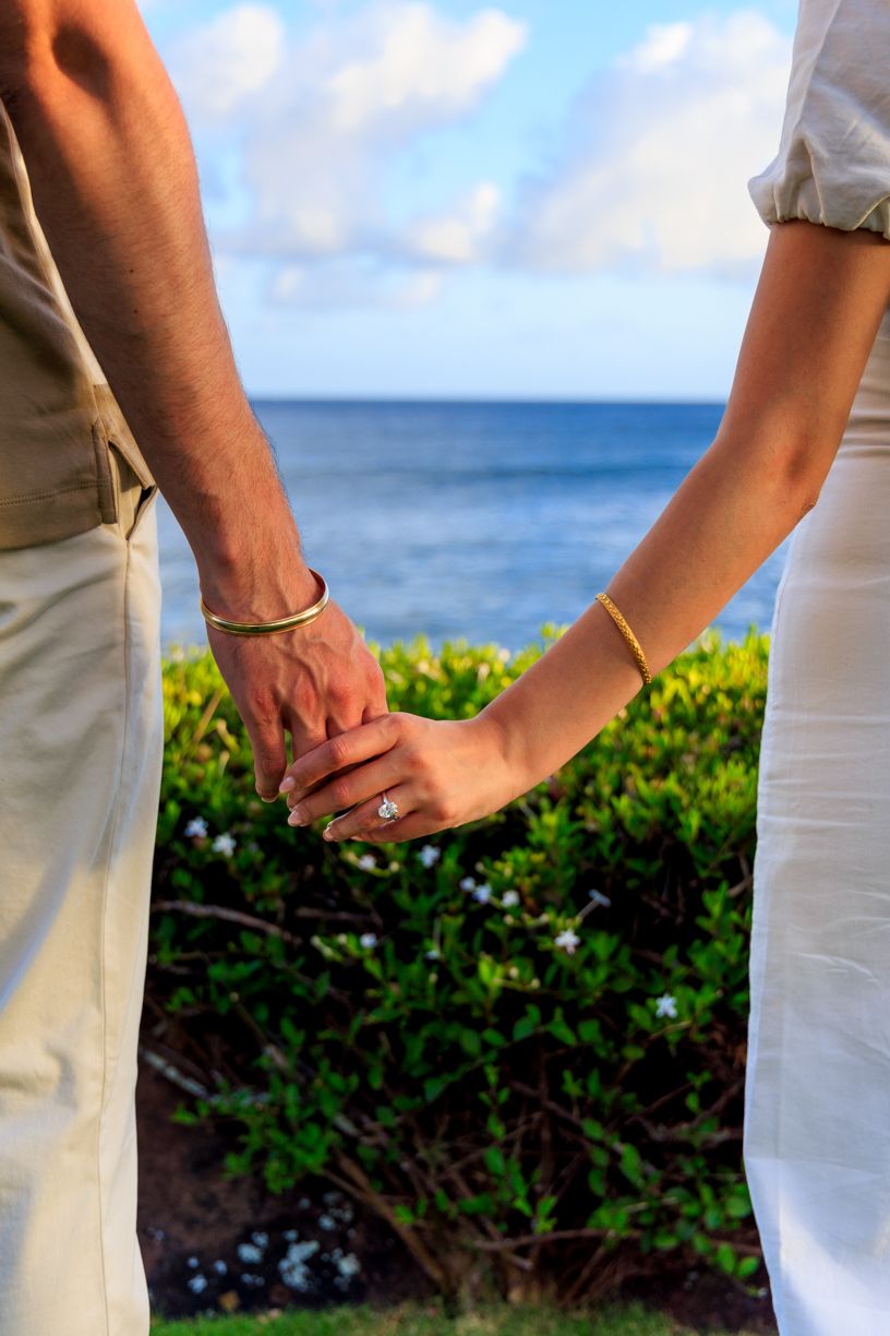Detail shot of couple's hands with engagement ring against ocean backdrop.