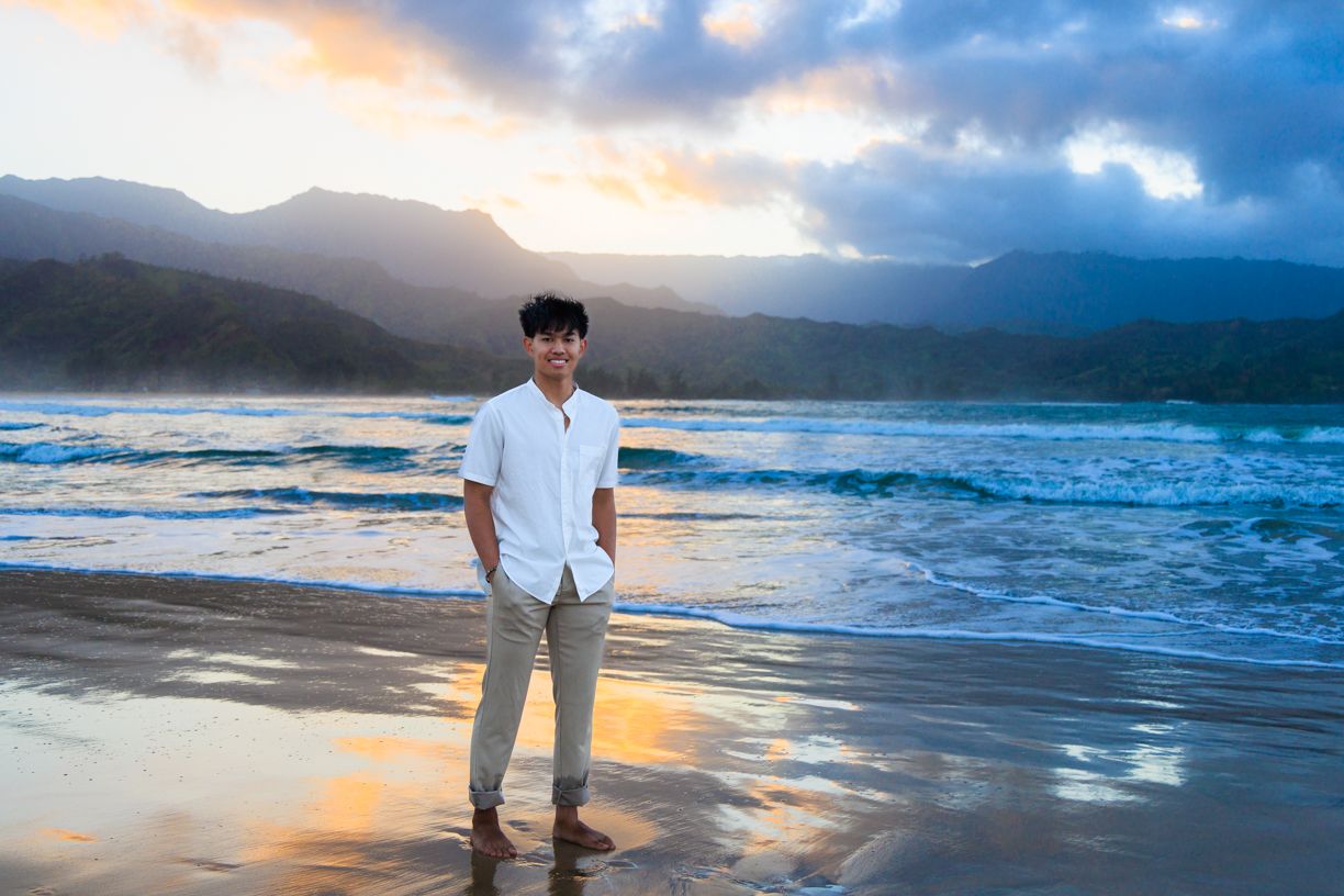 Man in white shirt and khaki pants standing on beach at sunset with mountains in background.