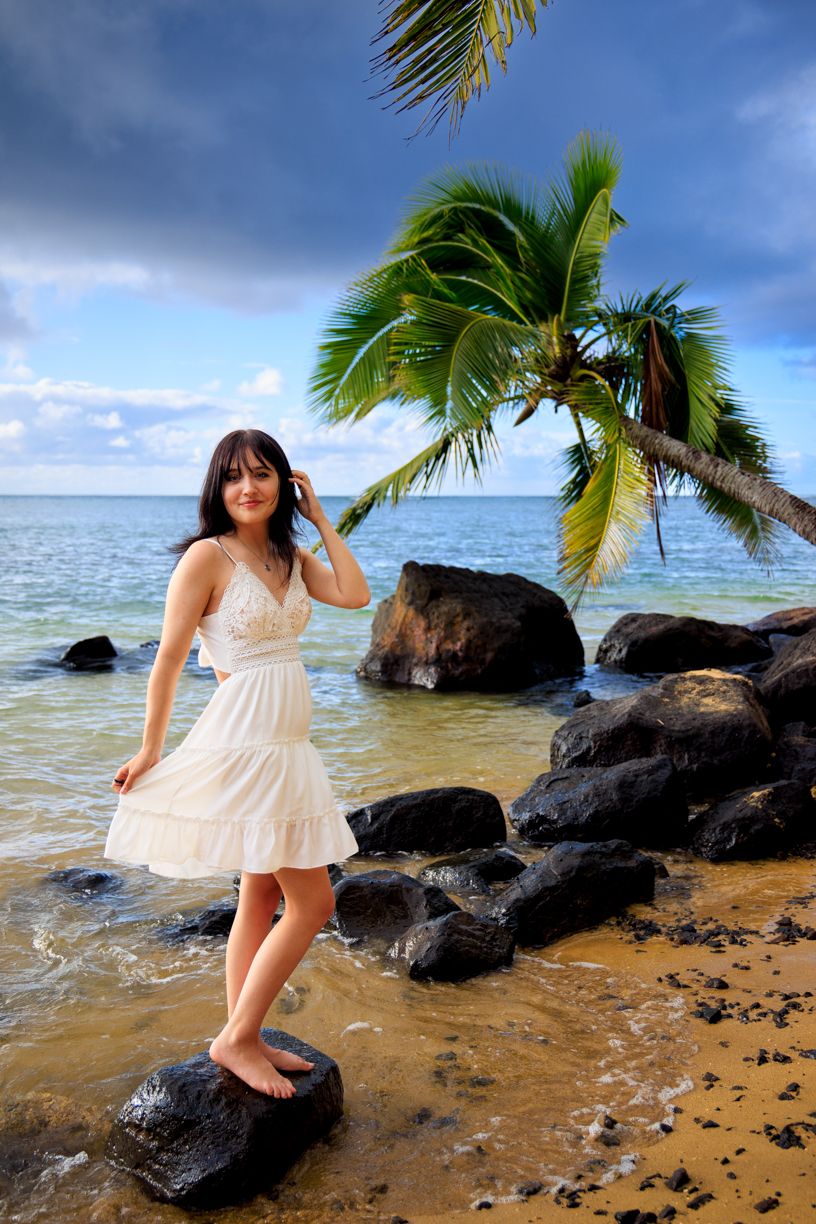 White dress flowing in the breeze on a tropical beach with palm trees and volcanic rocks at sunset.