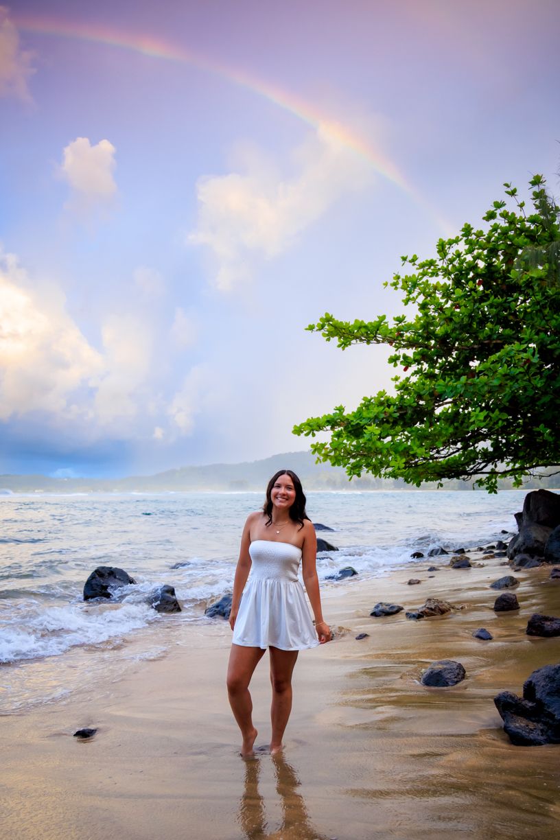 Scenic beach photoshoot with gentle waves, dark rocks, and lush greenery along the shoreline.