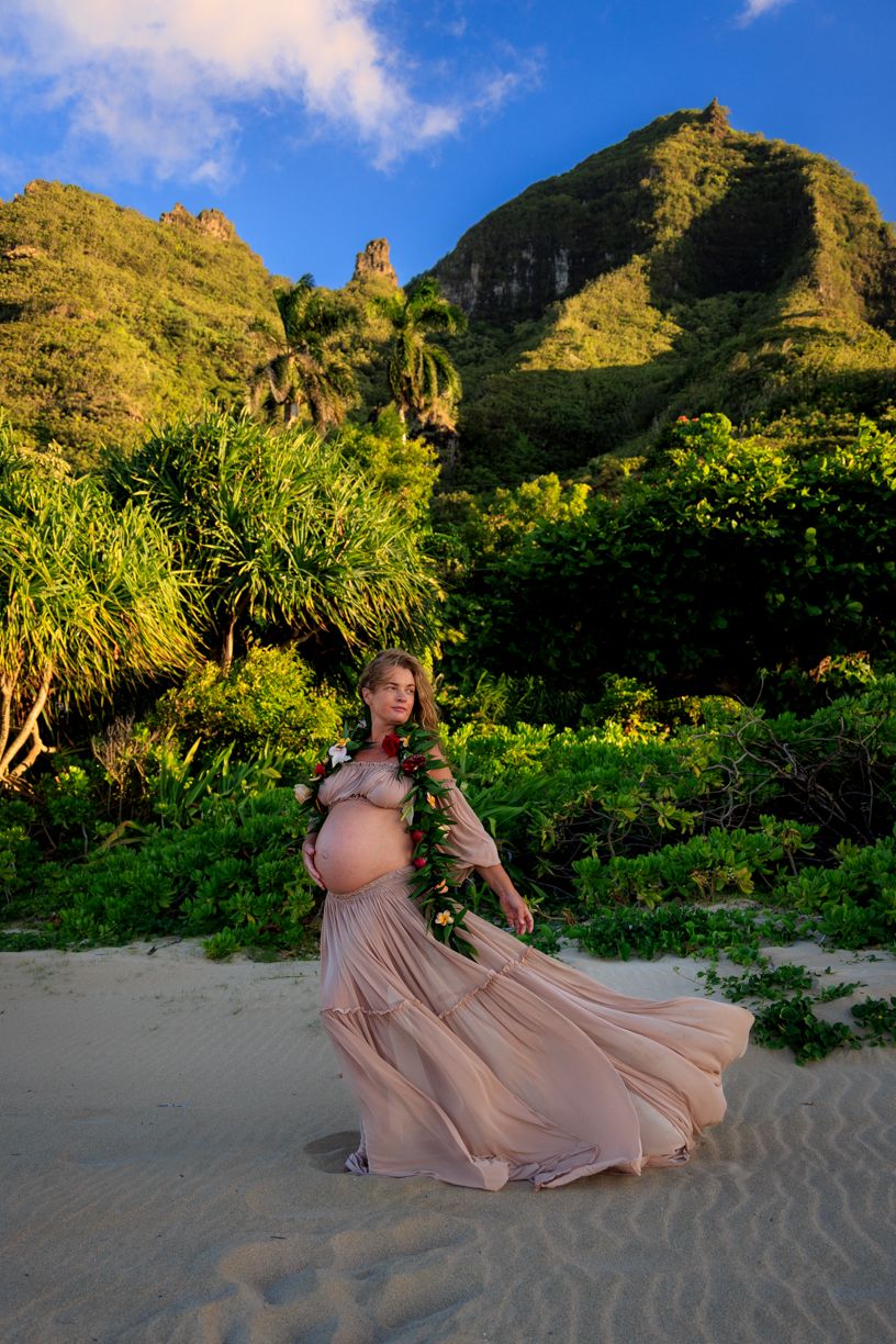 Maternity photo session on a tropical beach with mountains and lush greenery in the background.