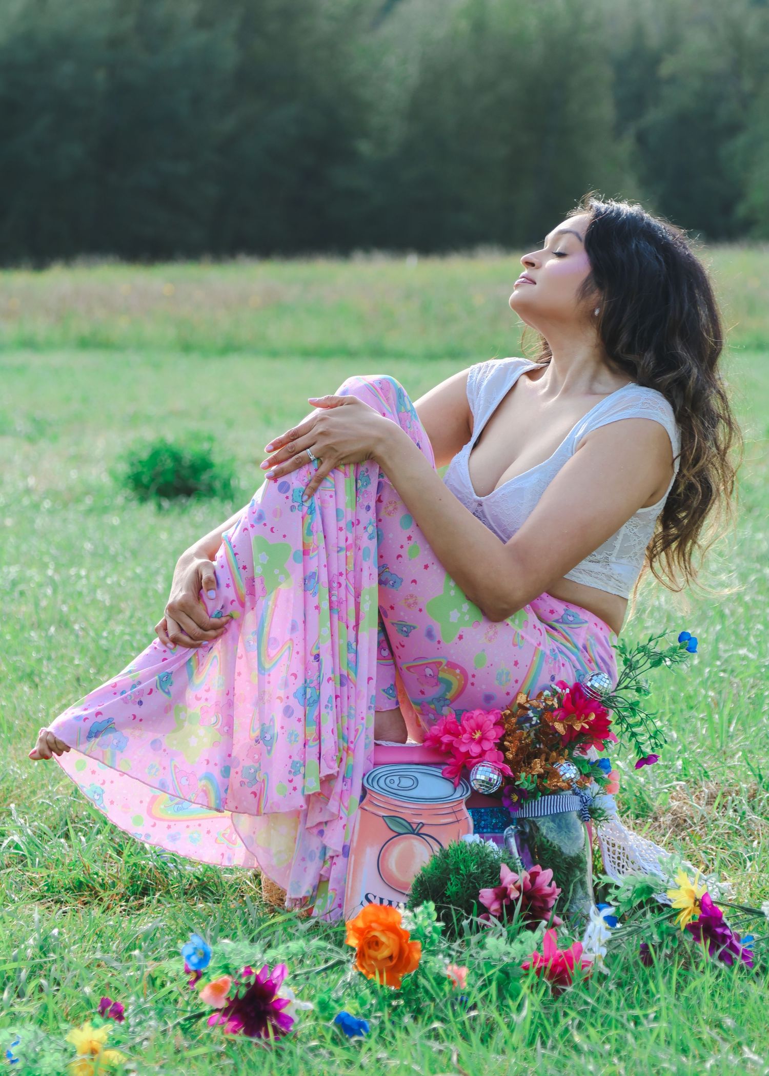 Woman in pink floral skirt and gray crop top sits in green meadow surrounded by colorful flowers.