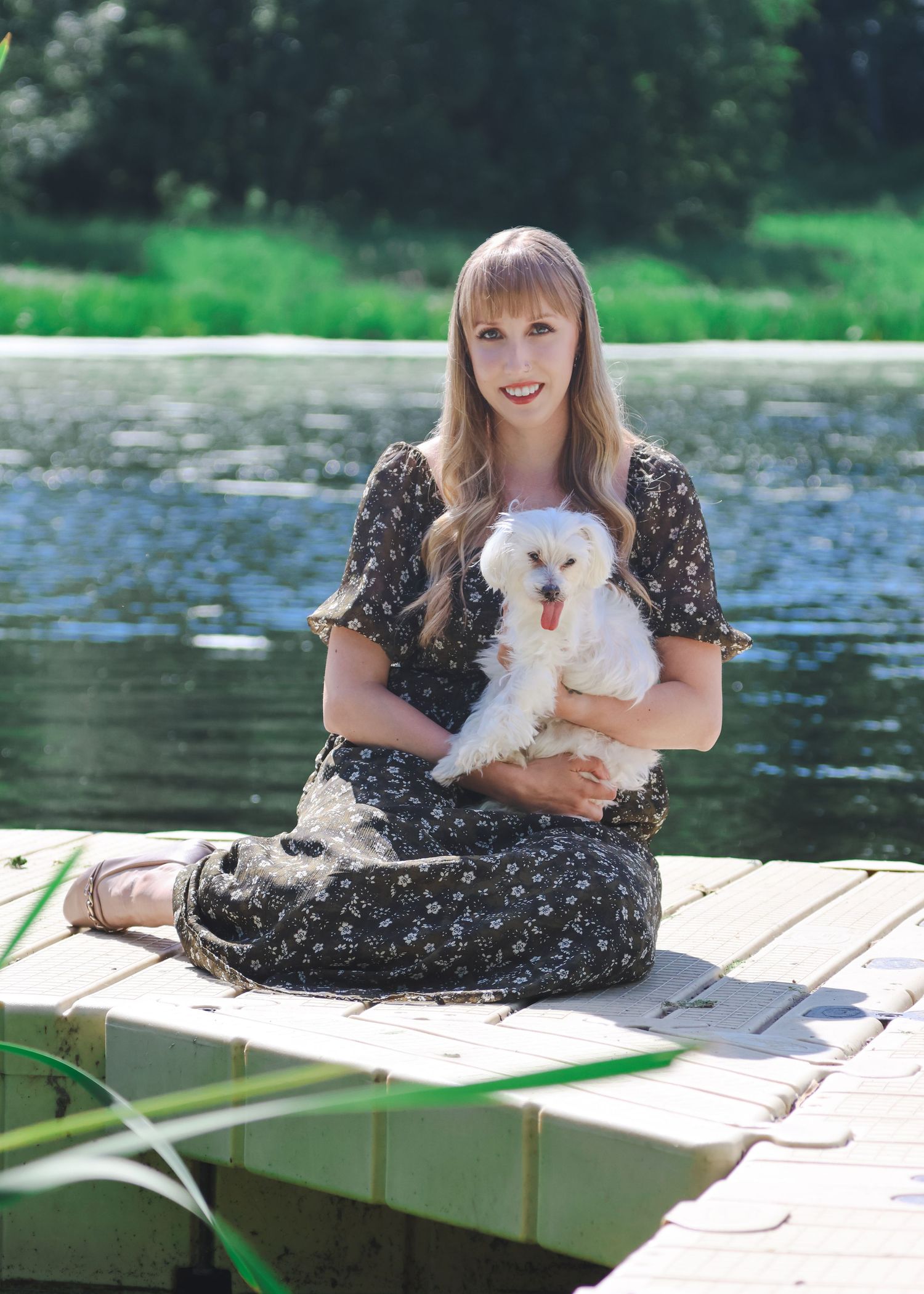Woman in floral dress sitting cross-legged on a dock holding a white fluffy dog near a serene lake.