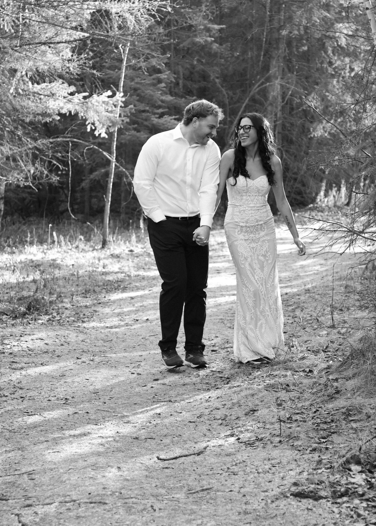 Black and white photo of newlyweds walking hand in hand along a sunlit forest trail.