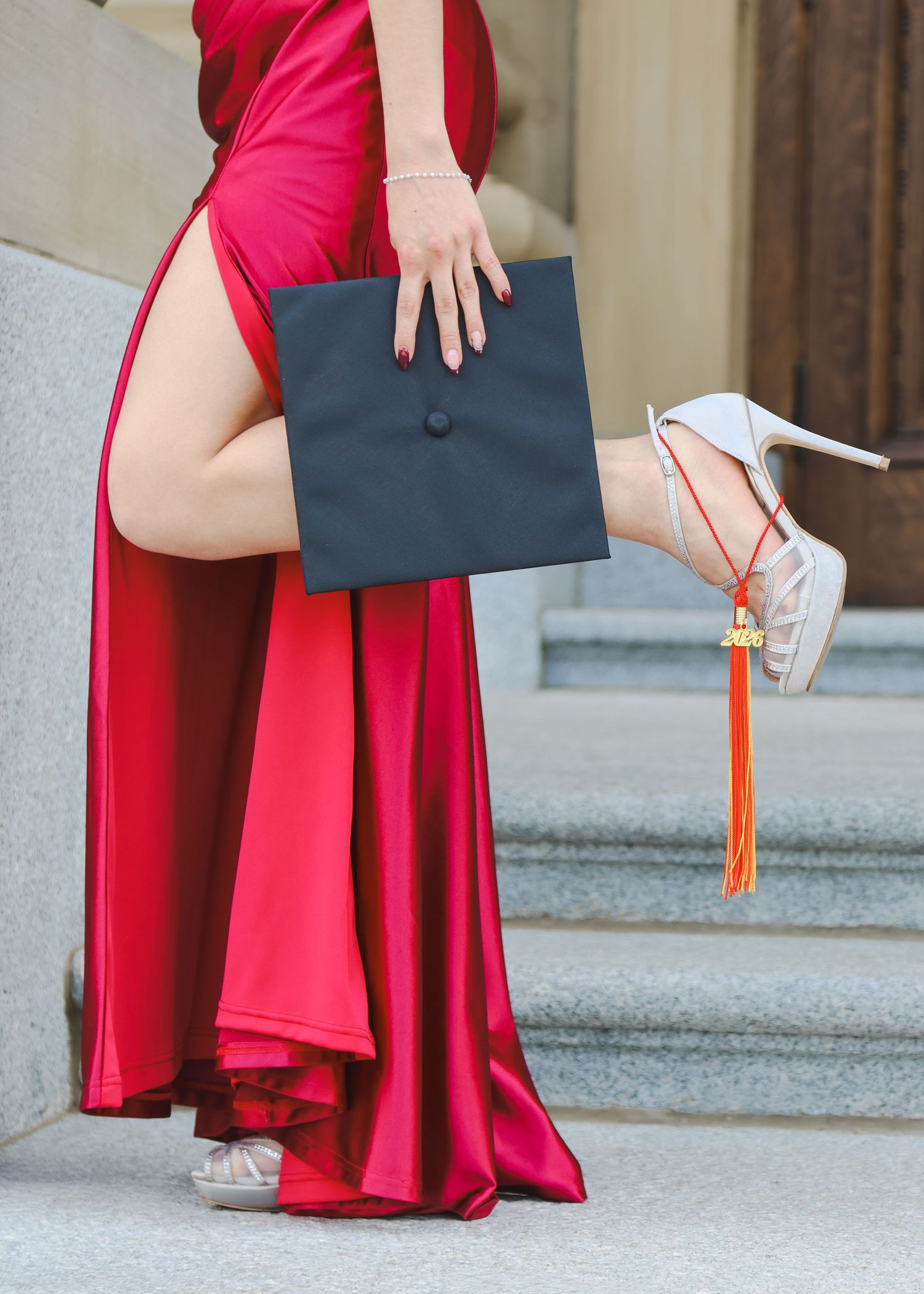 Female graduate in red formal gown holding black mortarboard cap while wearing white heels on steps.