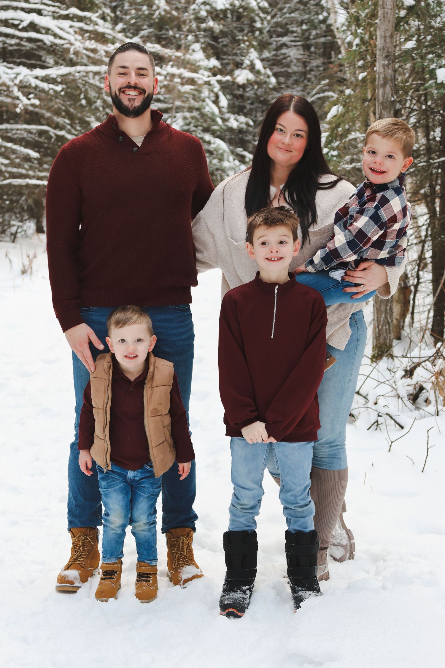 Family of five in burgundy and plaid stands together in snowy winter woodland setting.