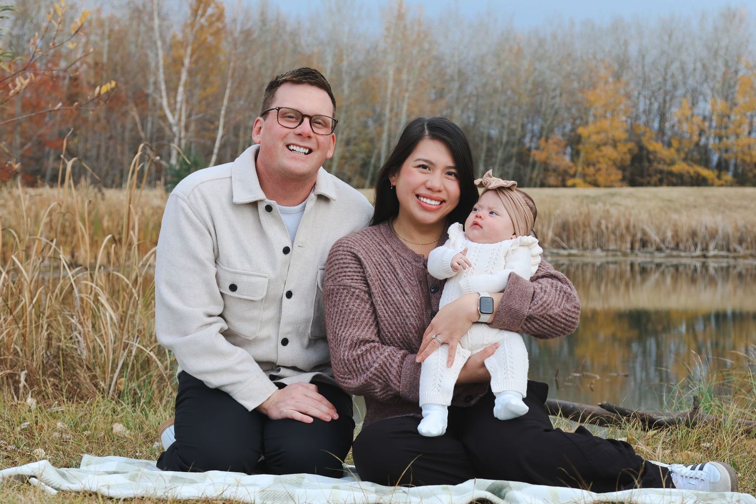 Smiling couple sits on blanket by pond holding baby in white outfit amid fall foliage.