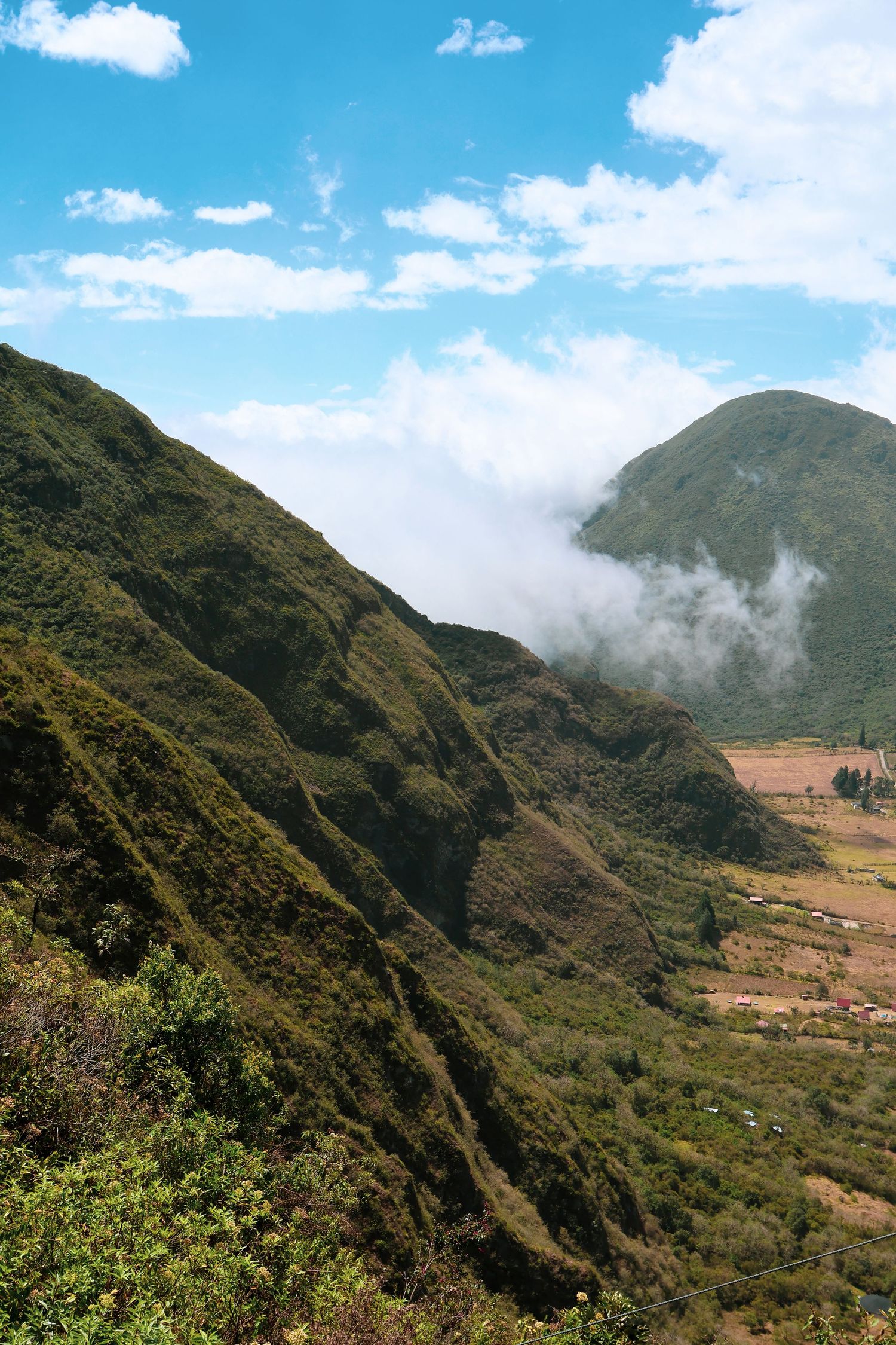Lush green mountain valley with low clouds, blue sky, and agricultural fields in a scenic Andean landscape.