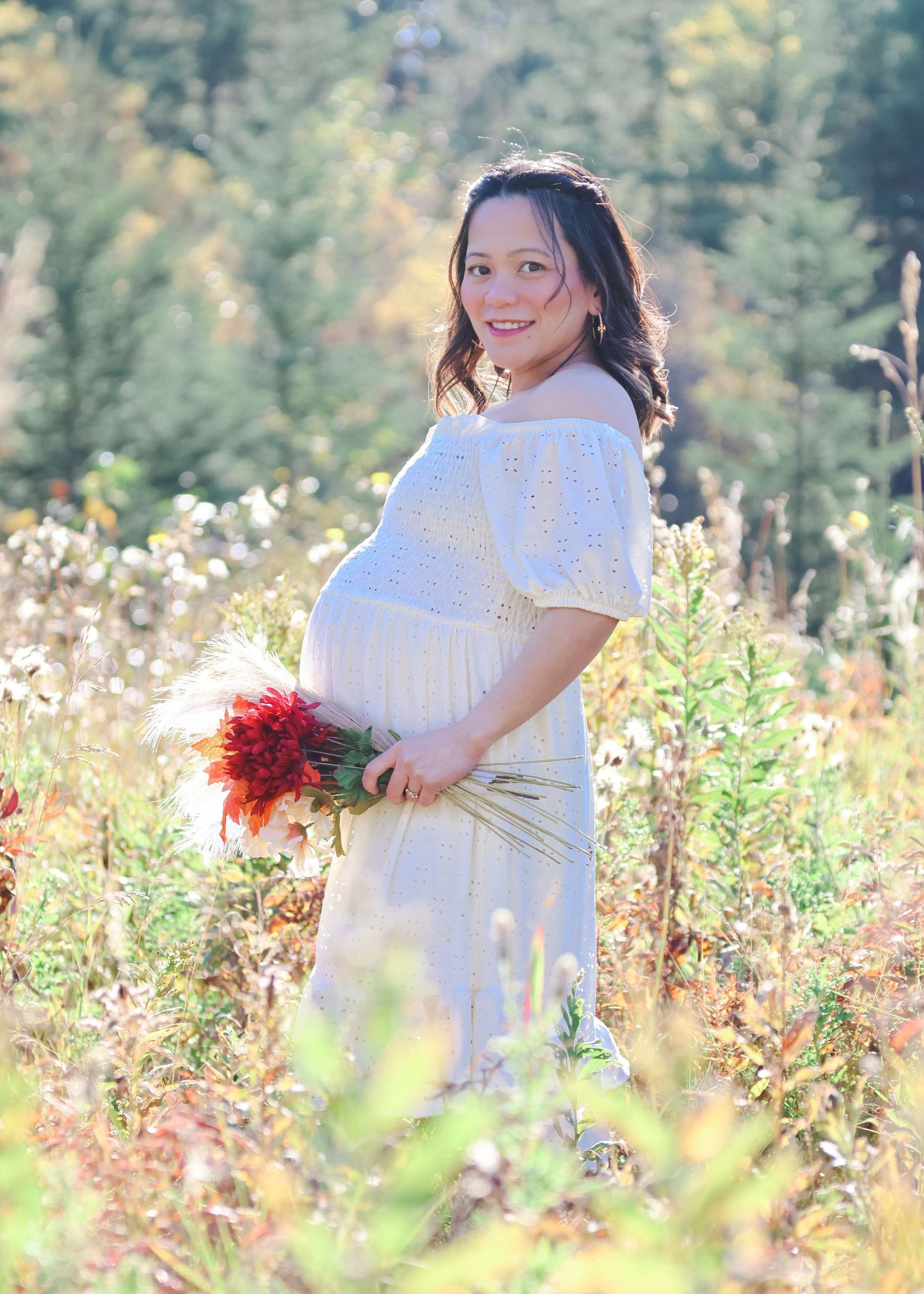 Pregnant woman in white lace dress holding red flowers in a sunlit autumn field.