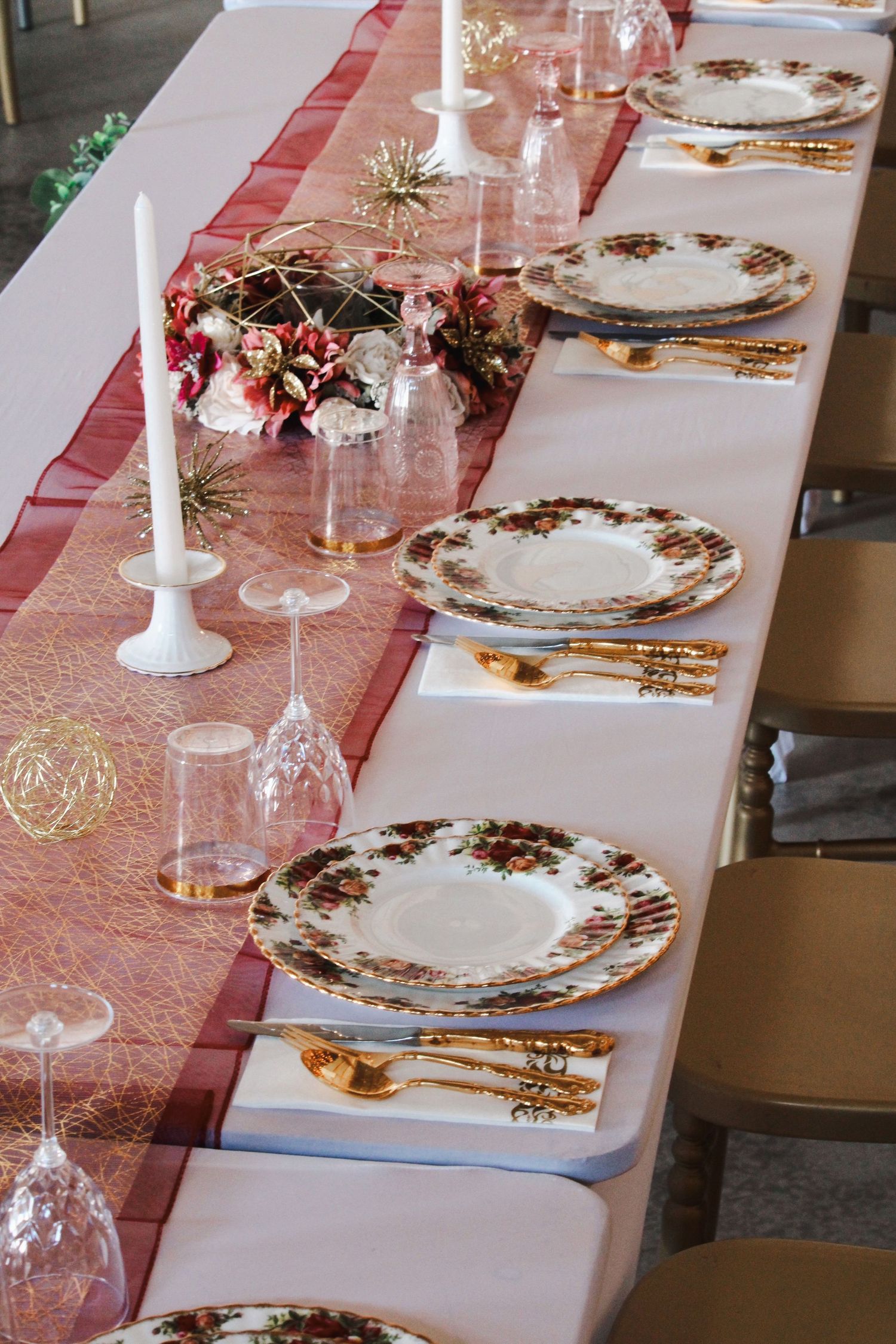 Elegant holiday table setting with floral china, gold cutlery, crystal glasses, and a red runner with candles.
