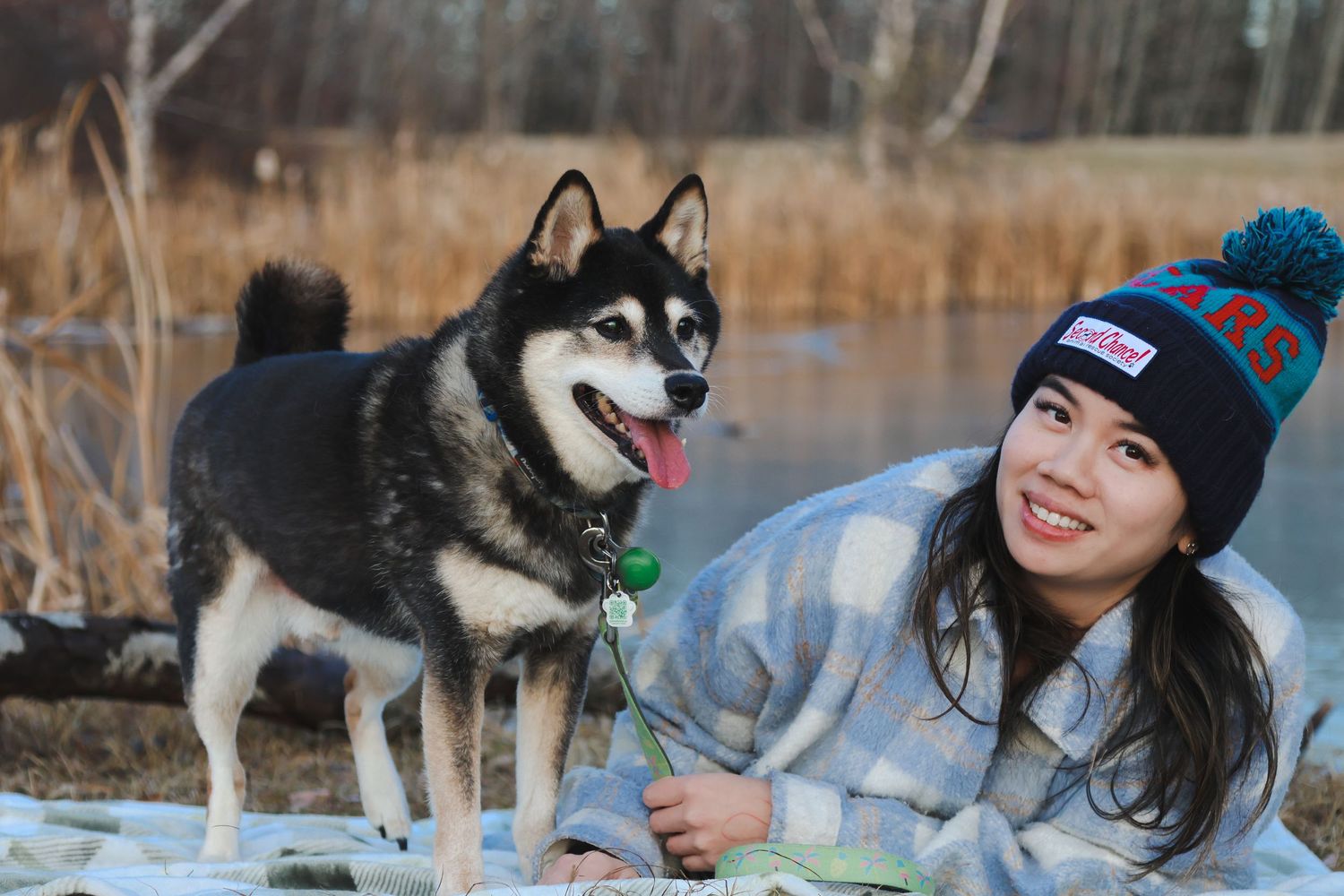Young woman in a pom-pom beanie smiles beside a black and white Husky dog near a winter lake.