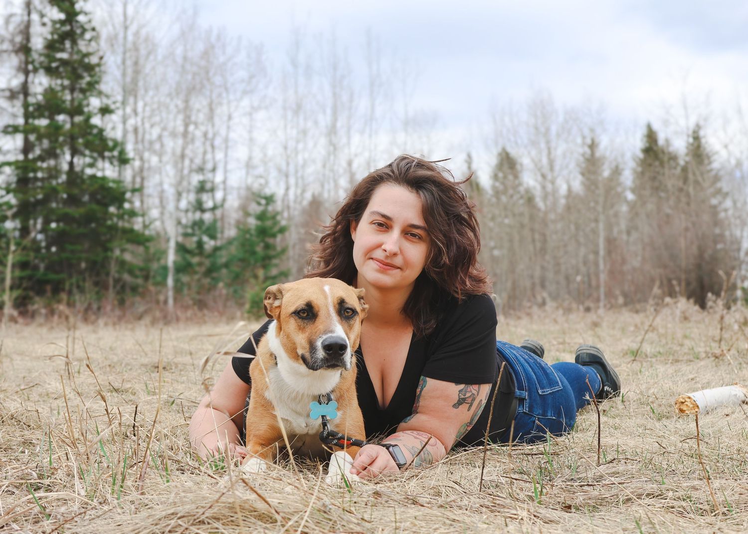 Woman lying in a field with her brown dog, surrounded by bare trees in a rural outdoor setting.
