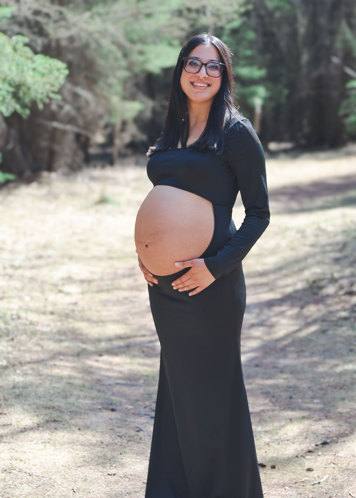 Pregnant woman in black crop top and maxi skirt cradling baby bump outdoors on forest path.