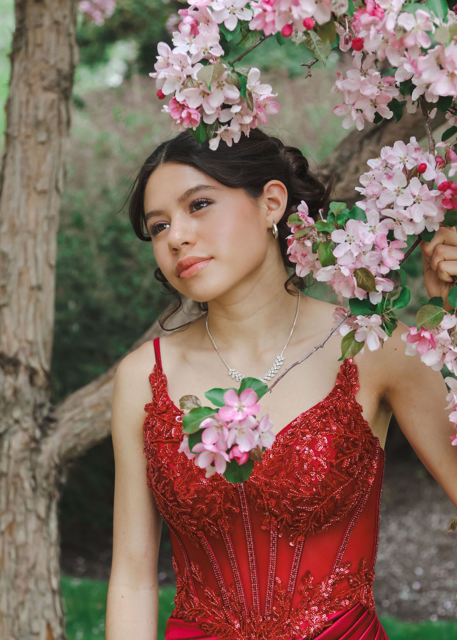 Young woman in red sequin formal gown posing near blooming pink flowers for graduation portraits.