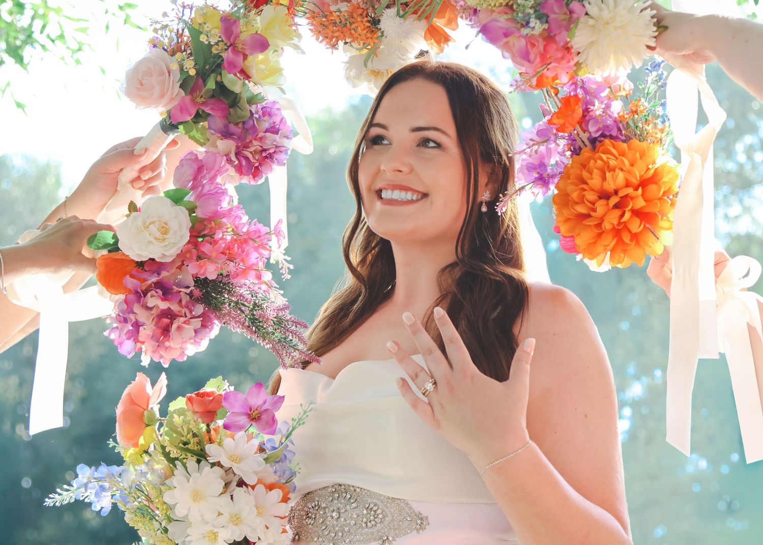 Smiling bride in white gown holding bouquet under colorful floral arch with pink and orange flowers.