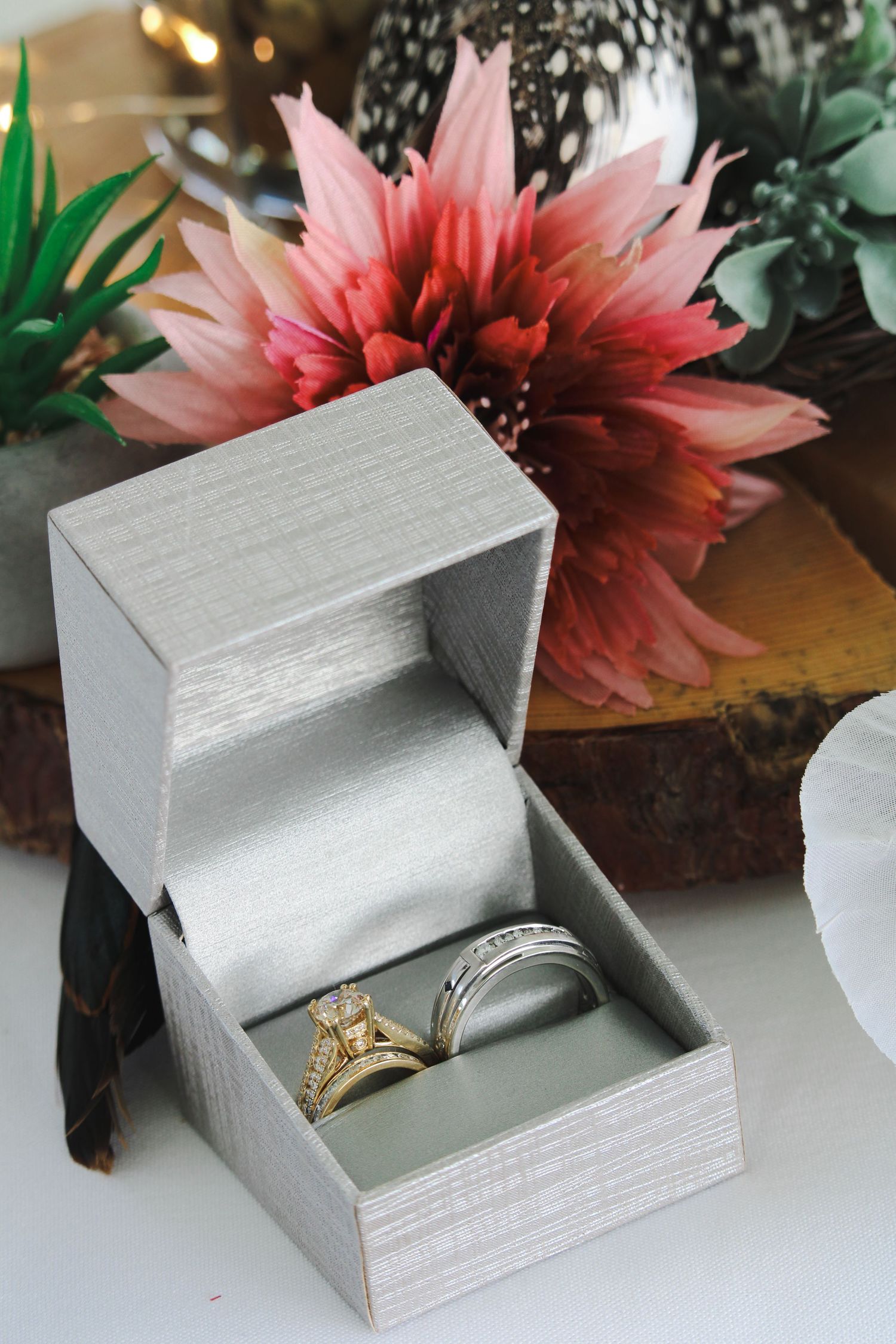 Gold engagement ring and silver wedding band displayed in a gray ring box with floral decor in the background.