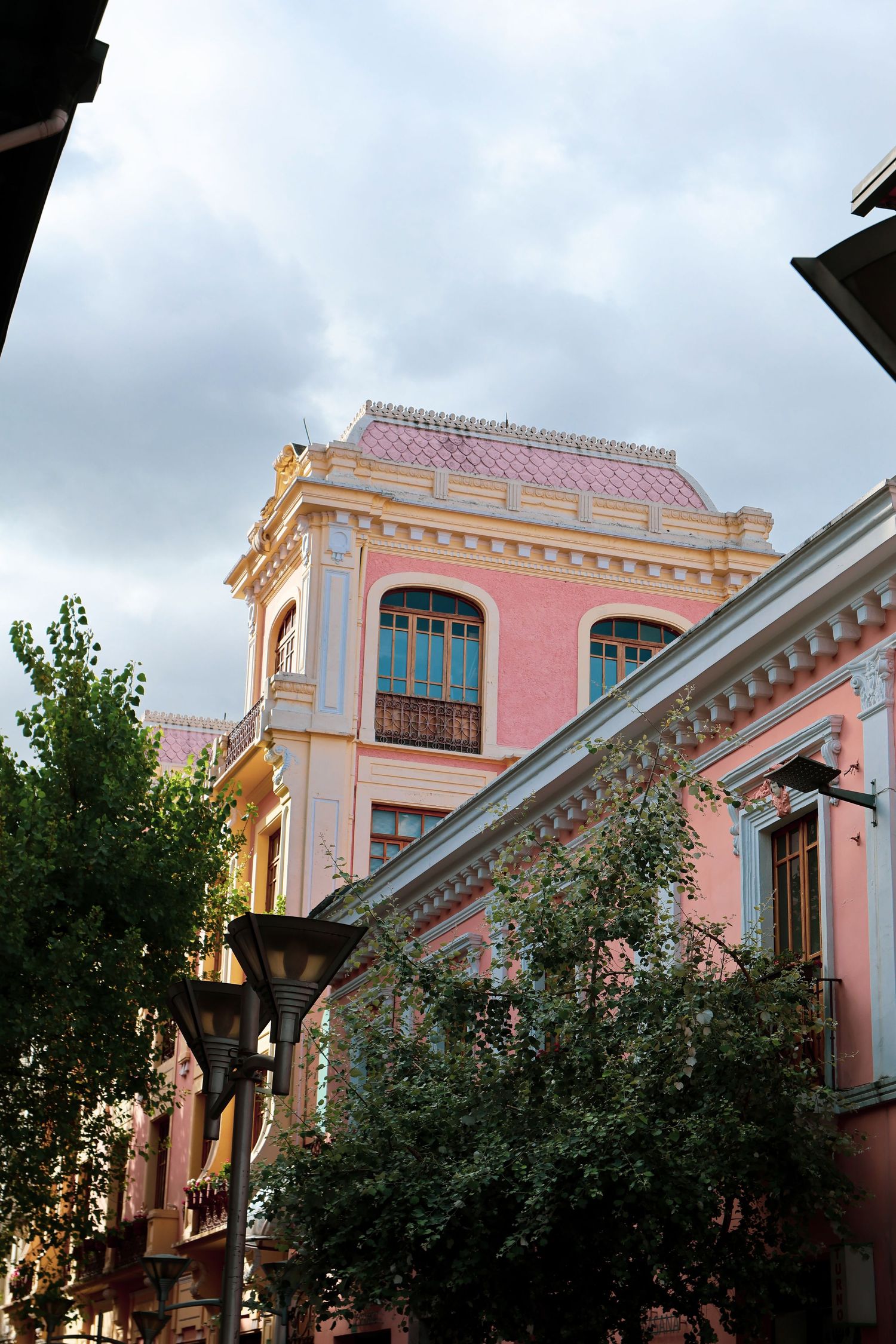Pink neoclassical building with ornate facade, blue windows, and lush green trees under a cloudy sky.
