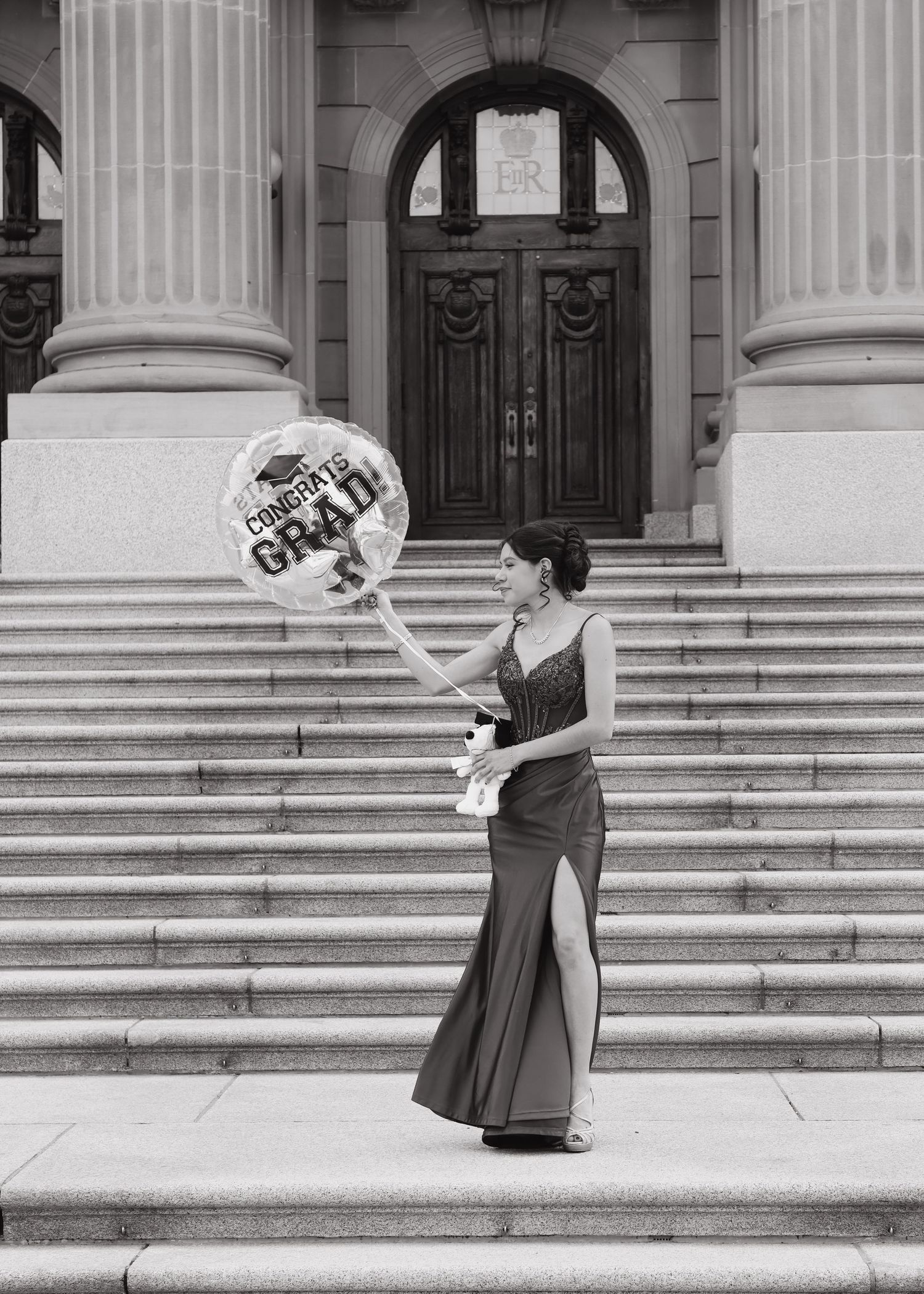 Black and white photo of woman in gown holding Congrats Grad balloon on steps.