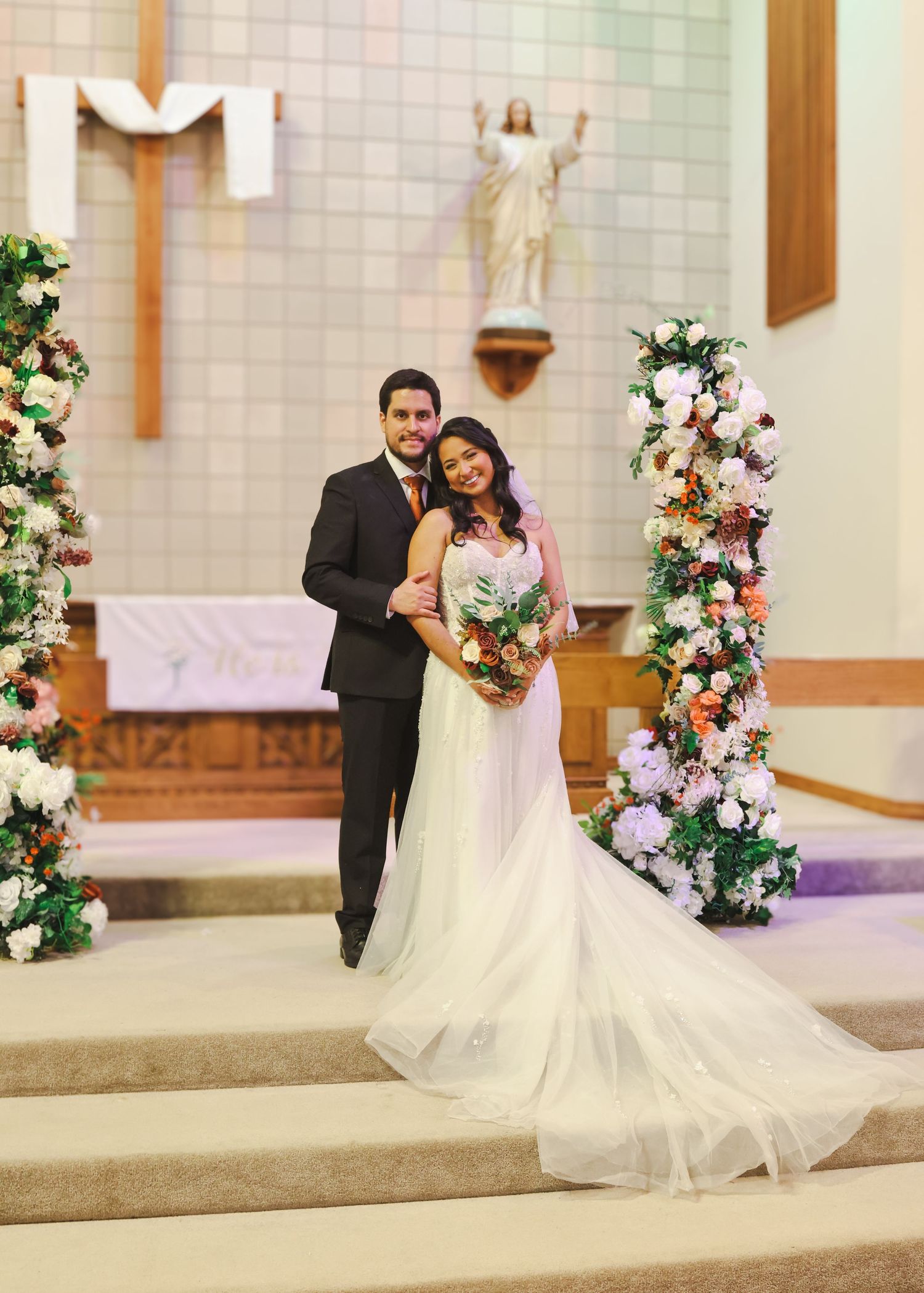 Bride and groom posing at church altar with floral columns and Jesus statue backdrop.