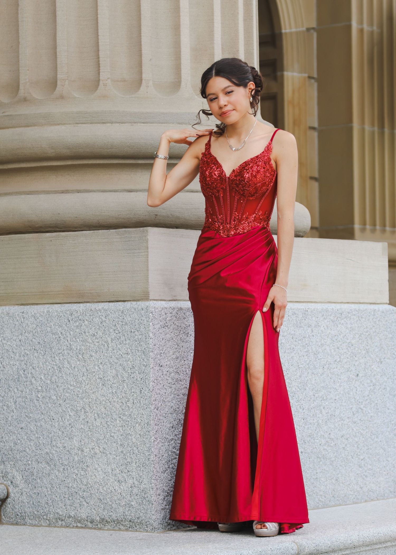 Young woman in elegant red sequin bodice gown with thigh slit posing by stone column.