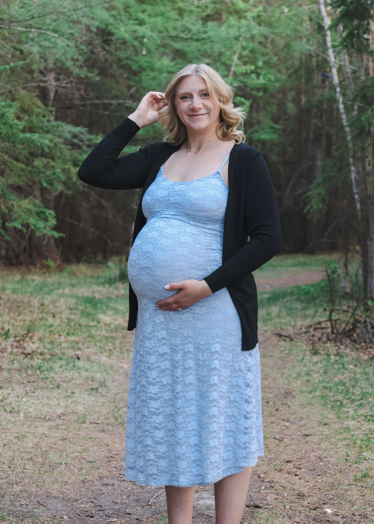 Pregnant blonde woman in light blue lace dress and black cardigan smiling outdoors in wooded setting.
