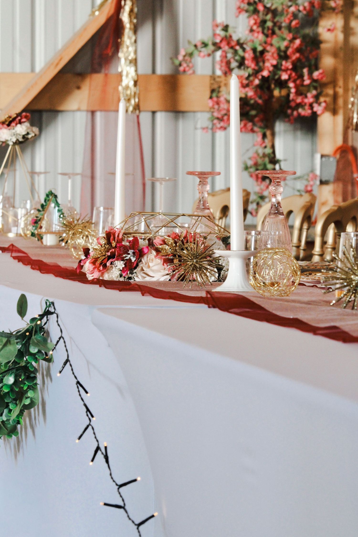 Elegant wedding head table with red runner, gold geometric decor, candles, and floral arrangements in a rustic barn venue.