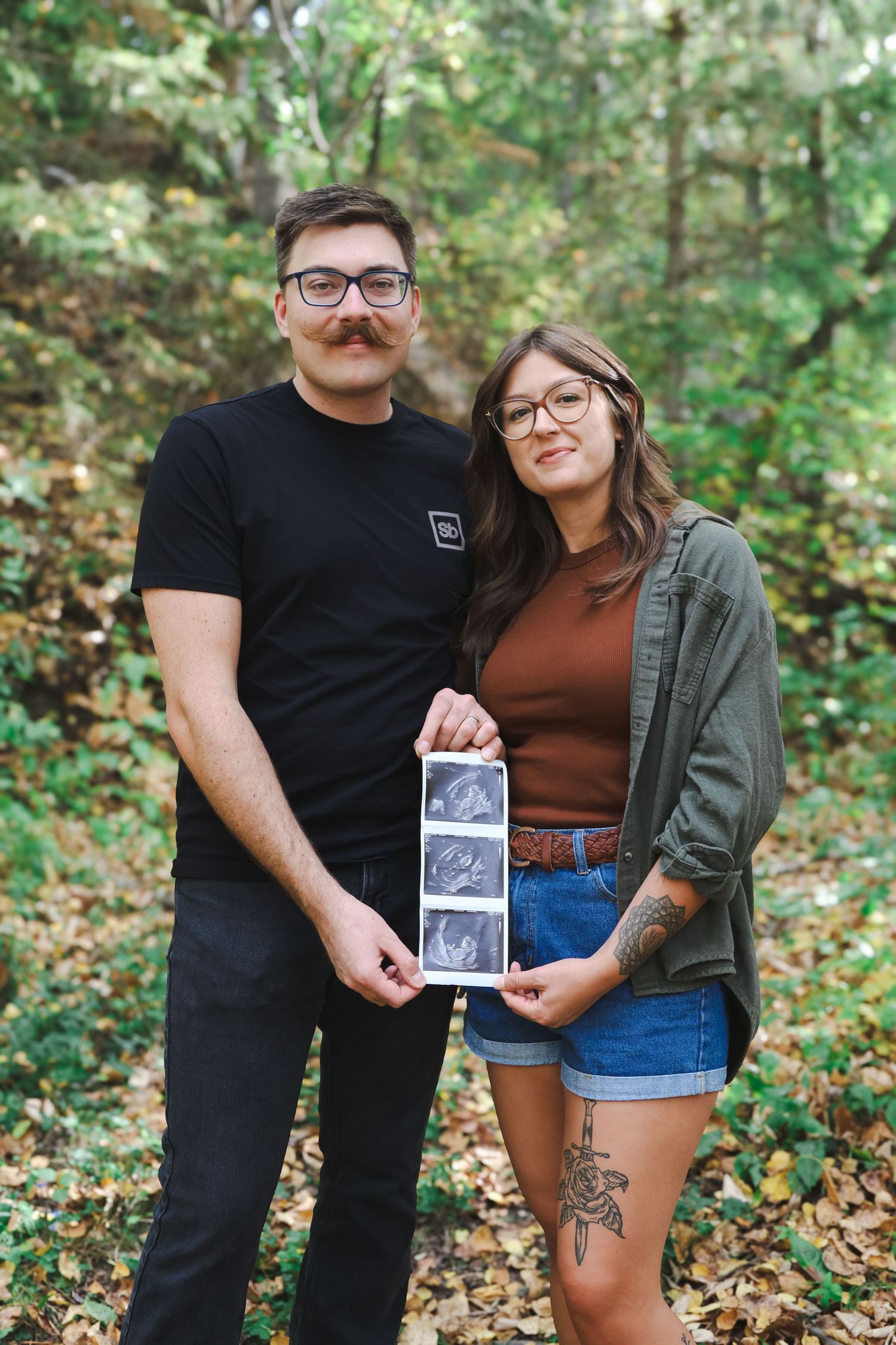 Couple holding ultrasound photos together in autumn forest for pregnancy announcement.