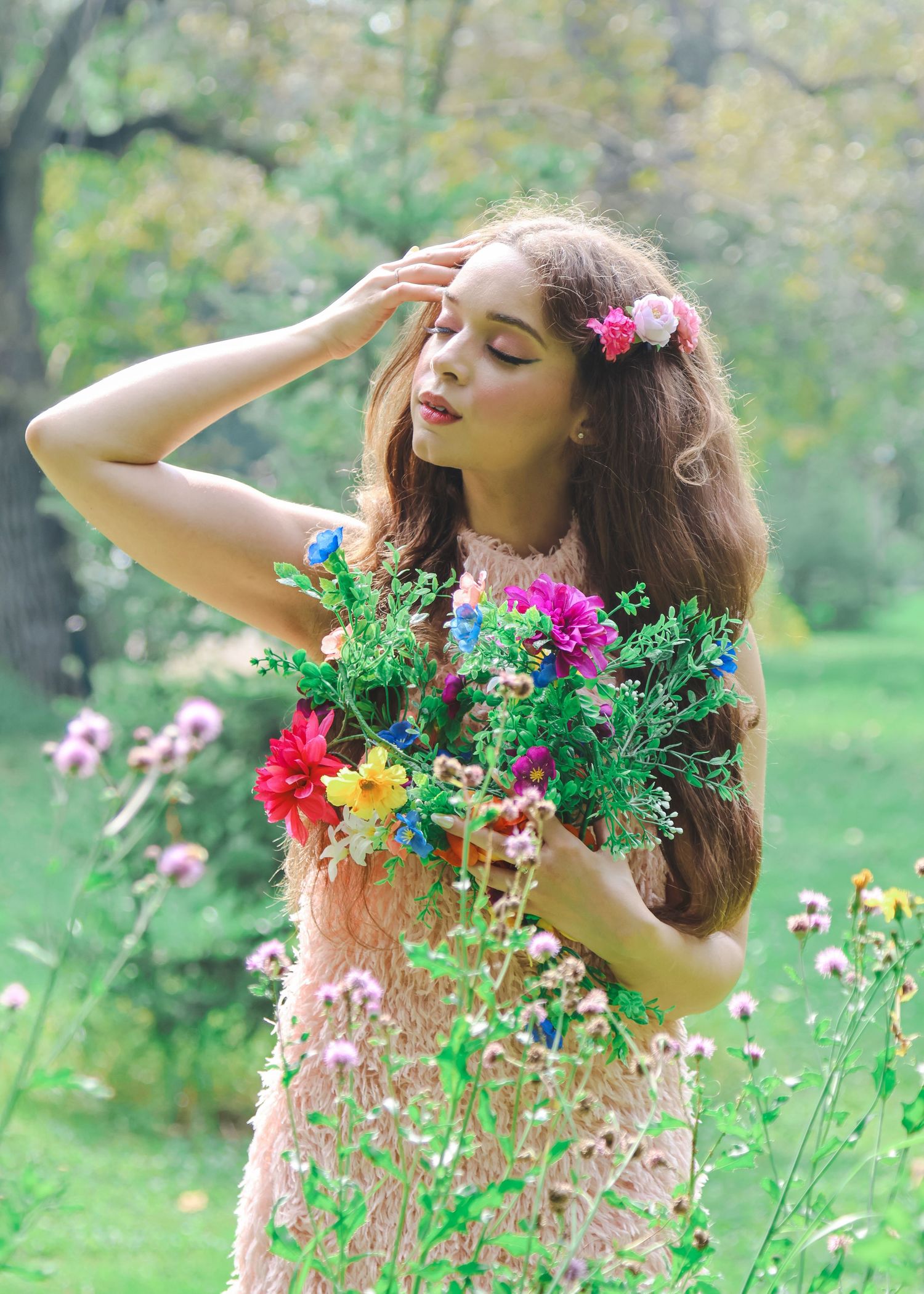 Young woman with flowers in her hair holds colorful bouquet in a lush green garden, wearing a pink dress.