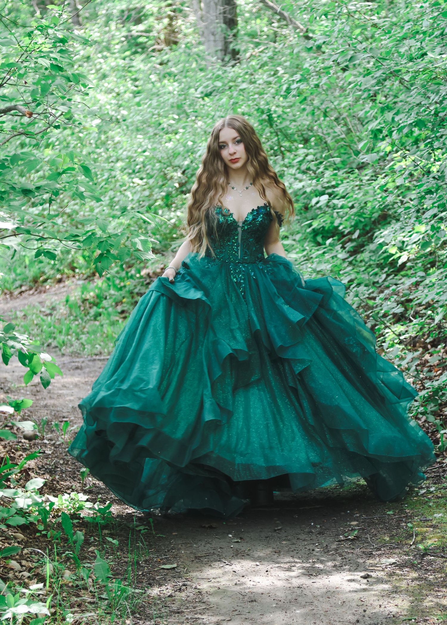 Young woman in a stunning emerald green ball gown walks along a forest path surrounded by trees.