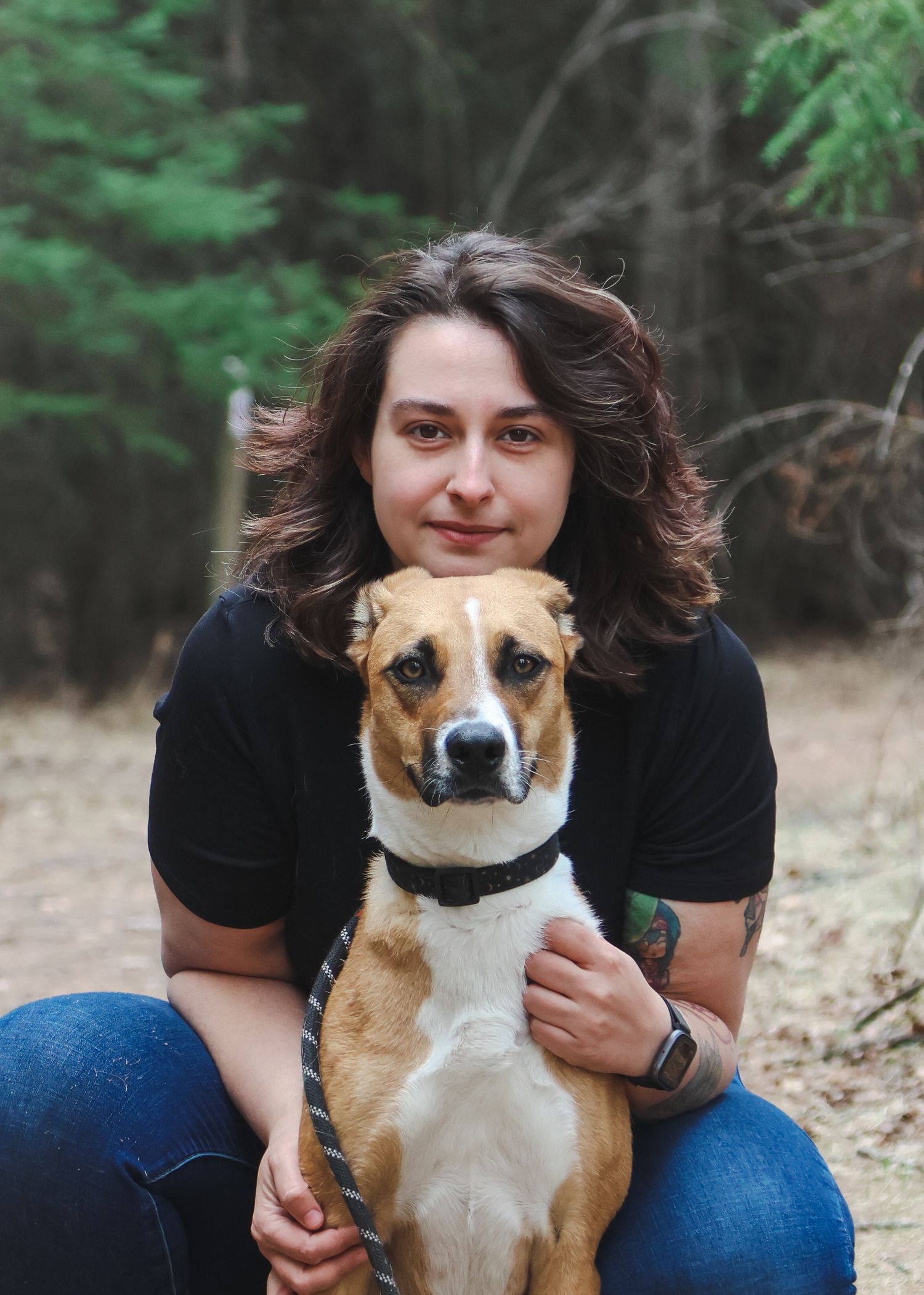 Woman with curly hair hugging a tan and white dog outdoors in a forest setting.