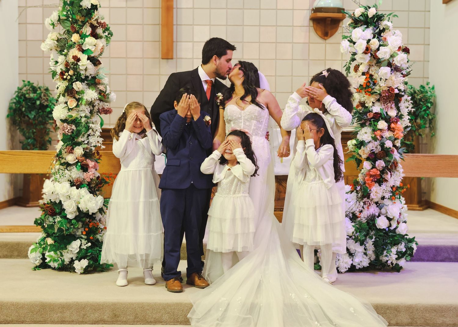 Wedding couple kissing at altar surrounded by flower girls covering their eyes in church.