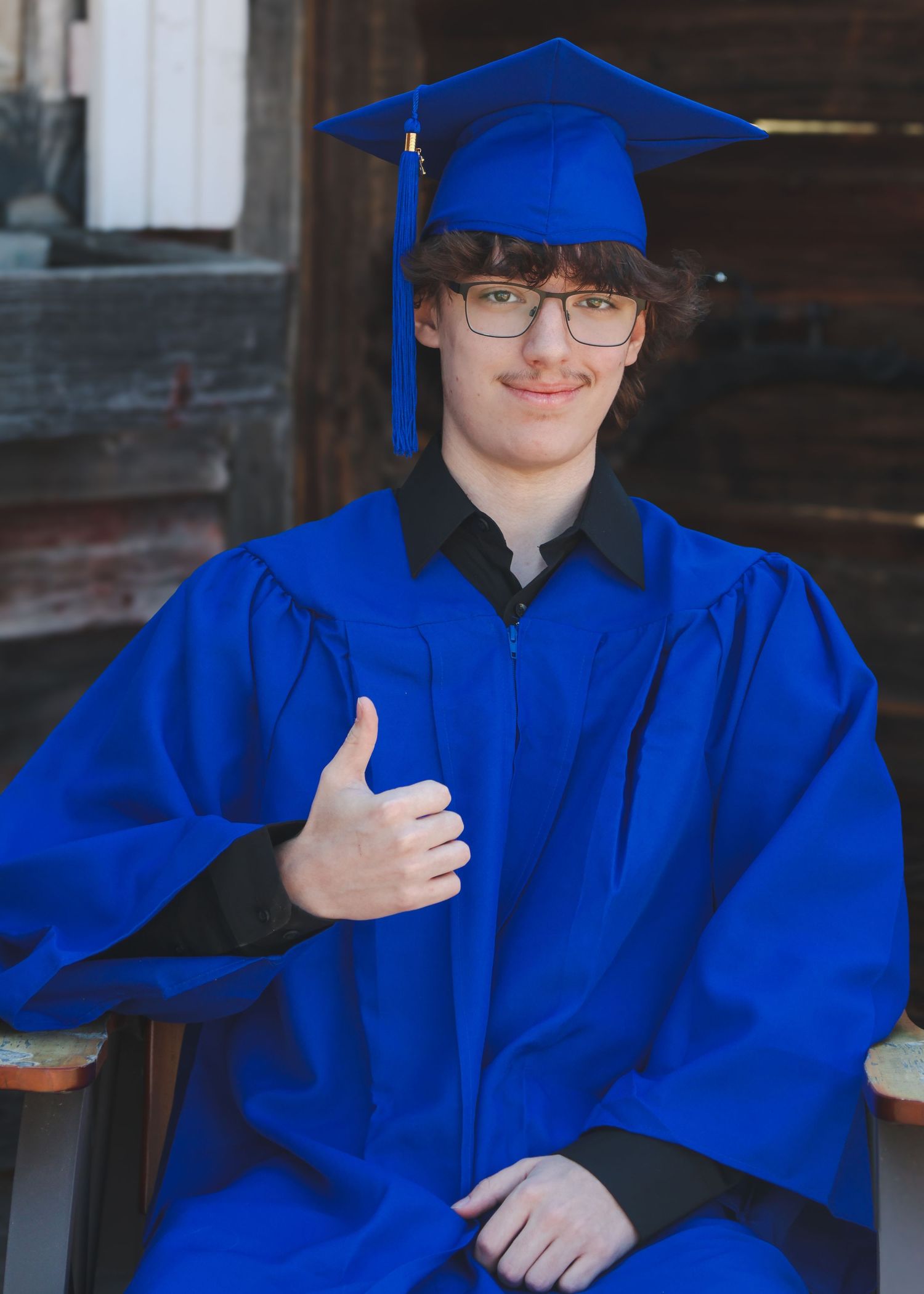 Smiling graduate in blue cap and gown giving thumbs up while seated for senior portrait photo.
