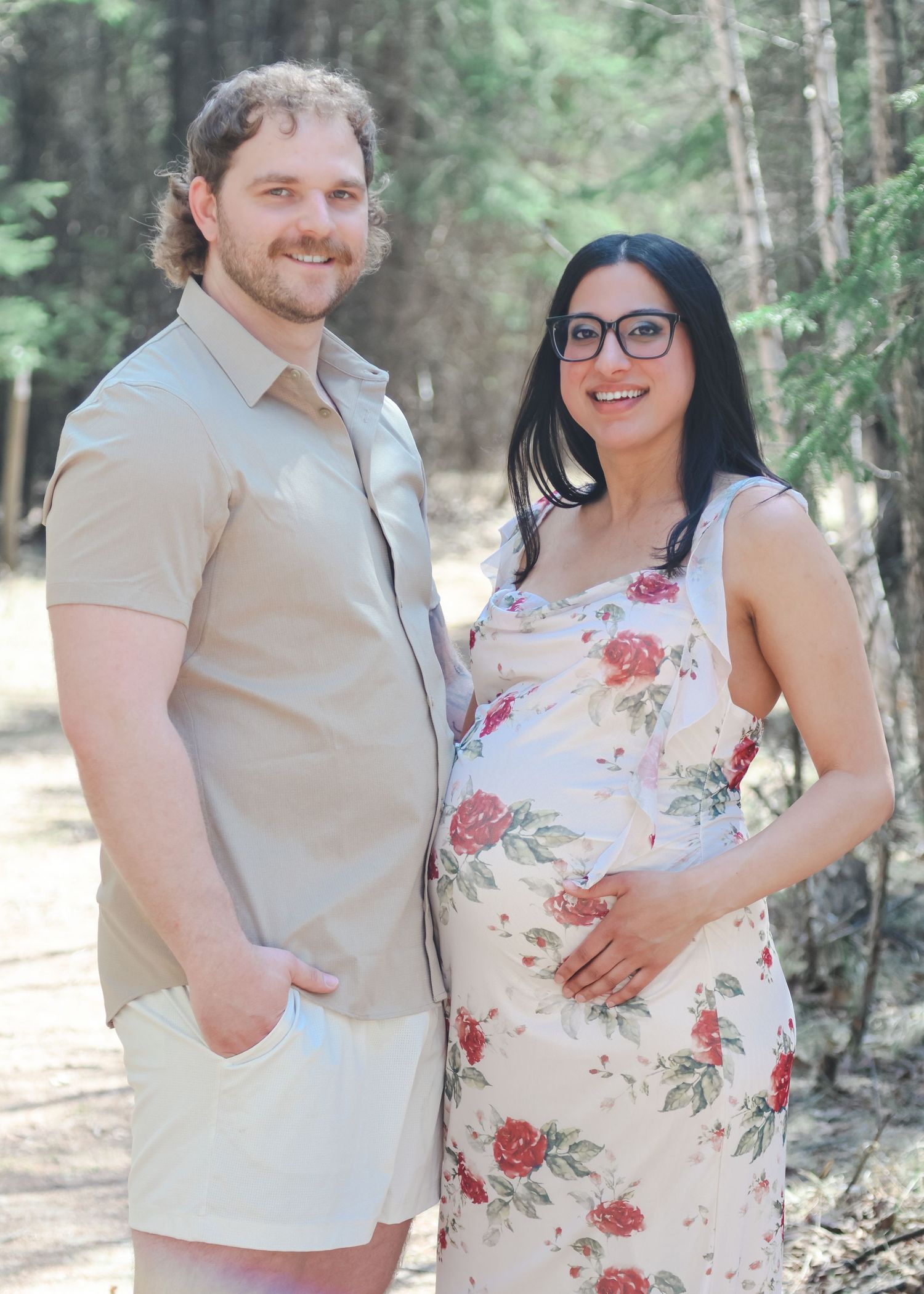 Smiling couple posing together outdoors, pregnant woman in floral dress holding baby bump beside partner.
