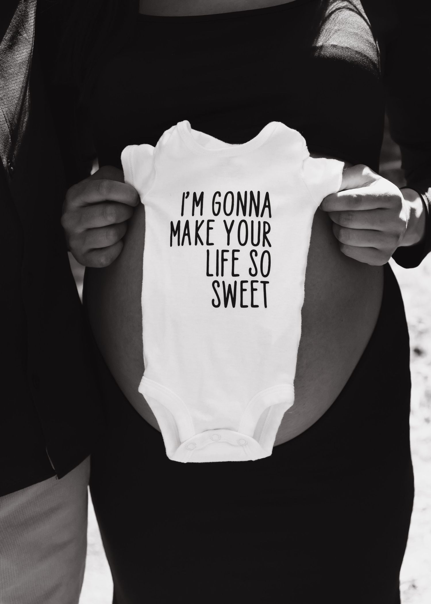 Pregnant woman holding white onesie reading I'm Gonna Make Your Life So Sweet.