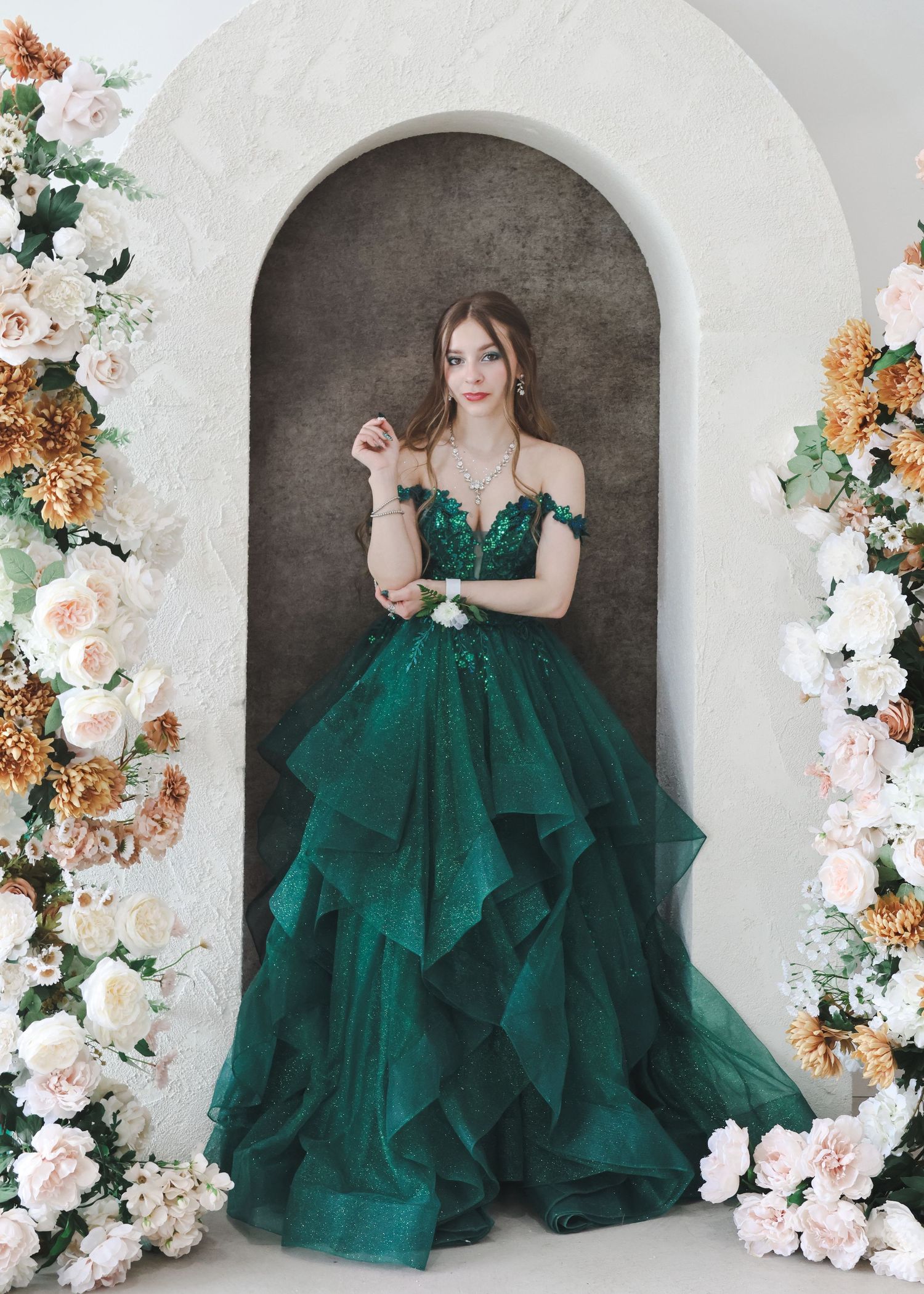 Woman in emerald green ball gown with lace bodice standing in floral arch doorway.