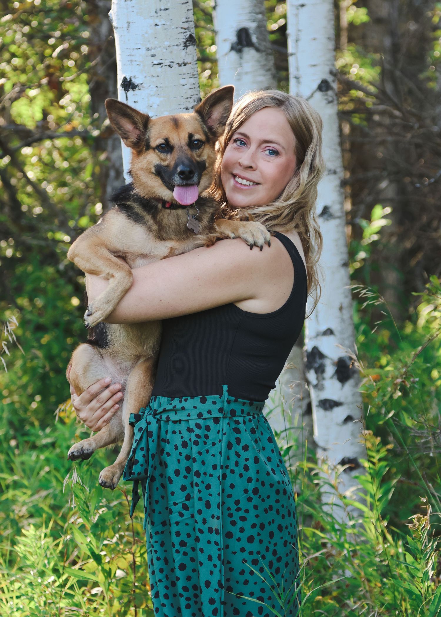Smiling blonde woman holding a tan German Shepherd mix dog outdoors near birch trees in a lush green forest setting.