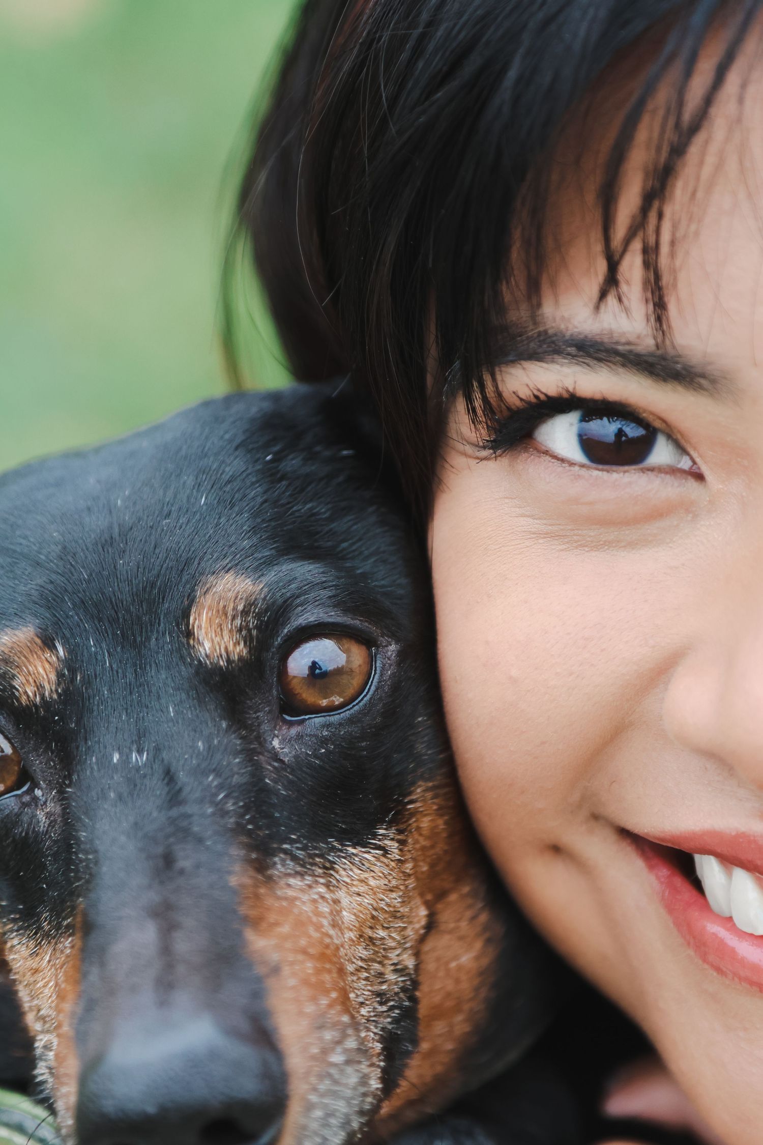 A smiling young woman with dark hair cheek-to-cheek with a black and tan Dachshund dog outdoors.