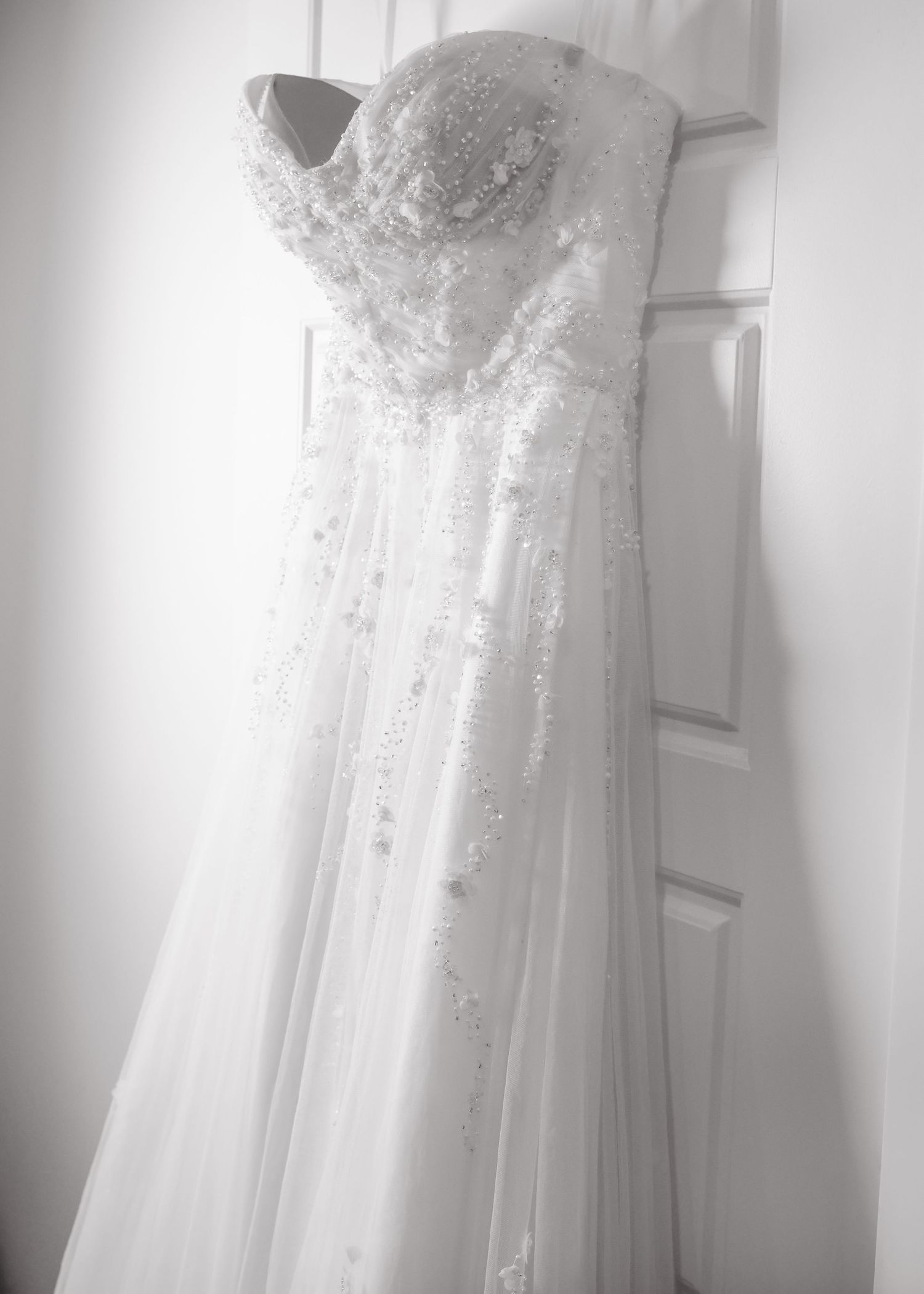 Black and white photo of an ornate beaded strapless wedding gown hanging on a door.