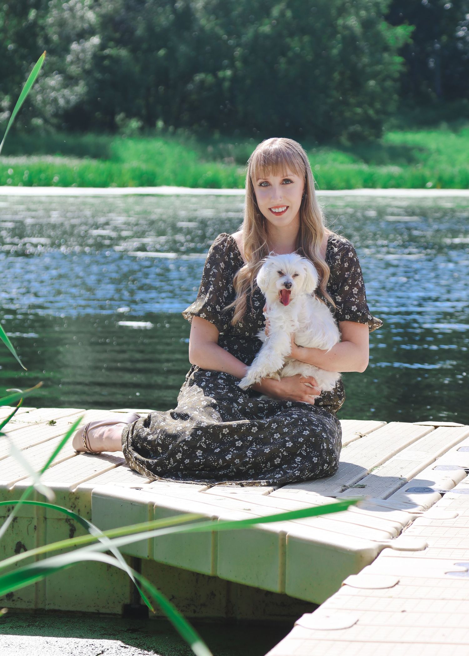Blonde woman in floral dress sitting on a lakeside dock holding a white fluffy dog outdoors.
