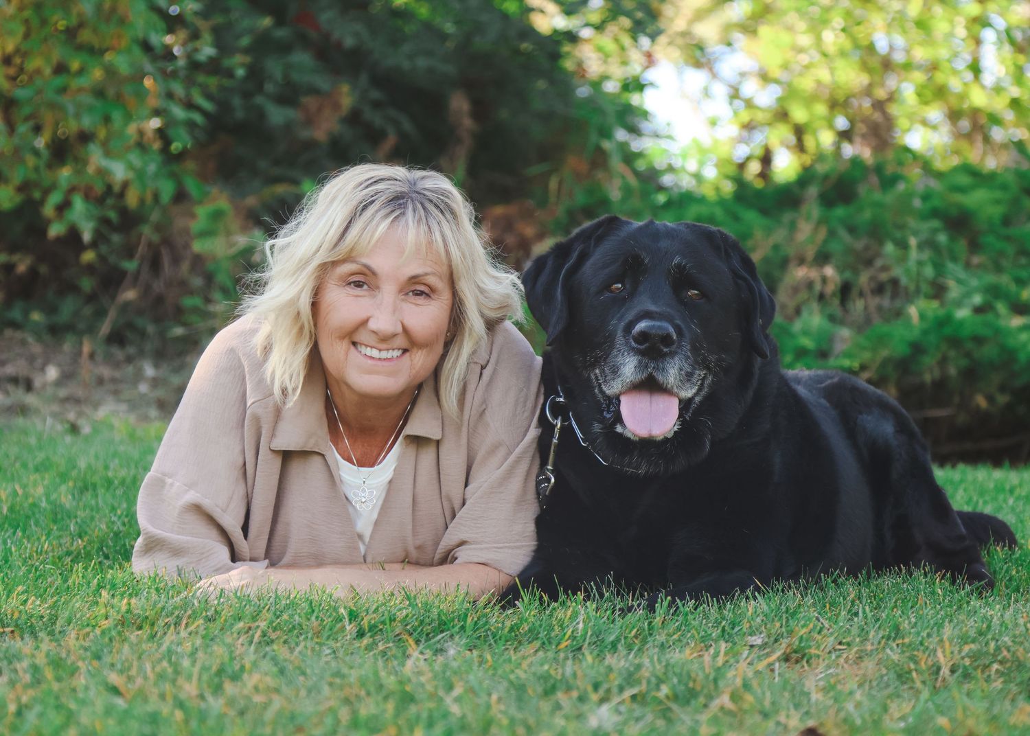 Smiling blonde woman lying on green grass beside a black Labrador Retriever dog outdoors.