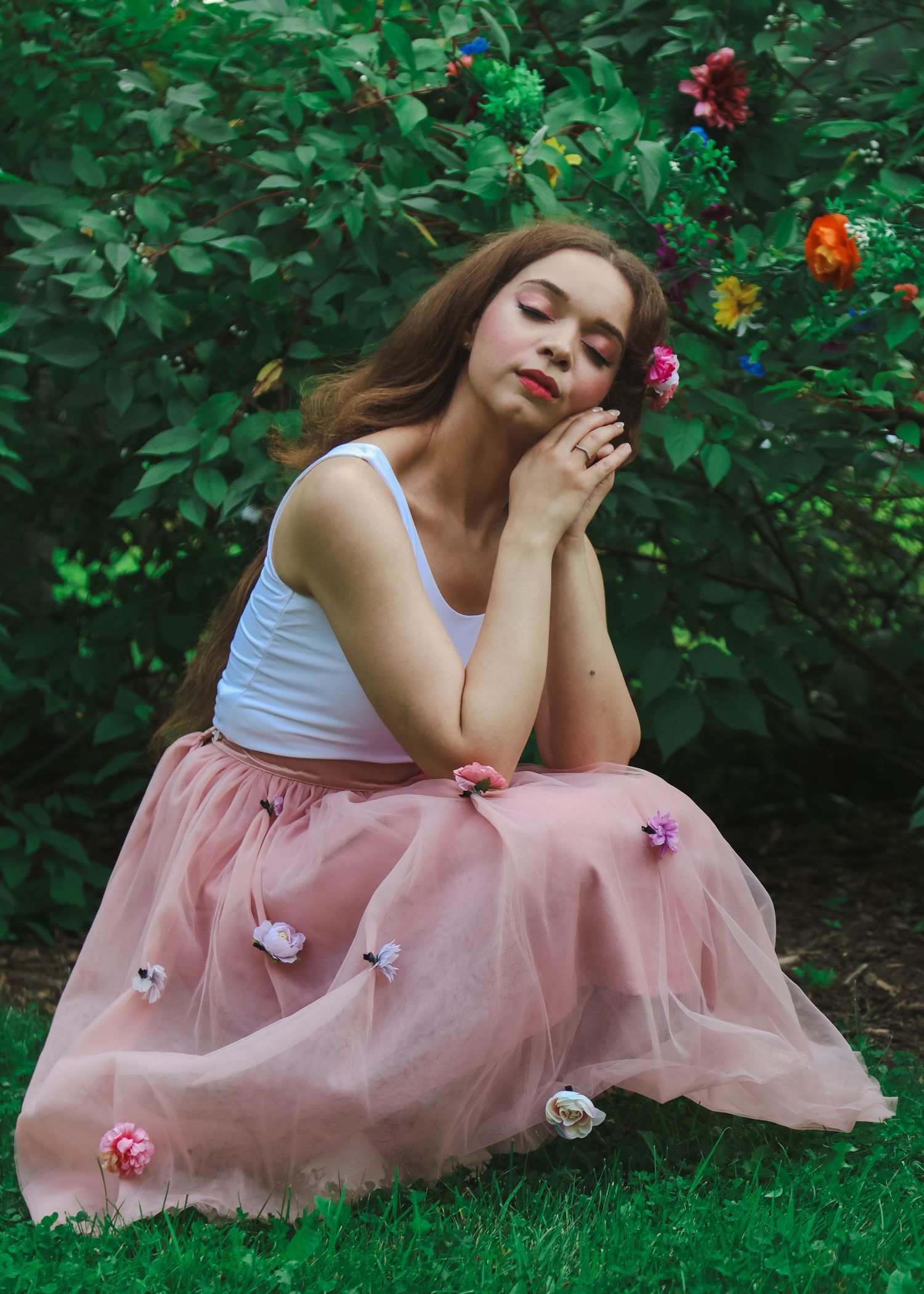 Young woman in white top and pink floral tulle skirt sitting peacefully in a lush green garden with colorful flowers.
