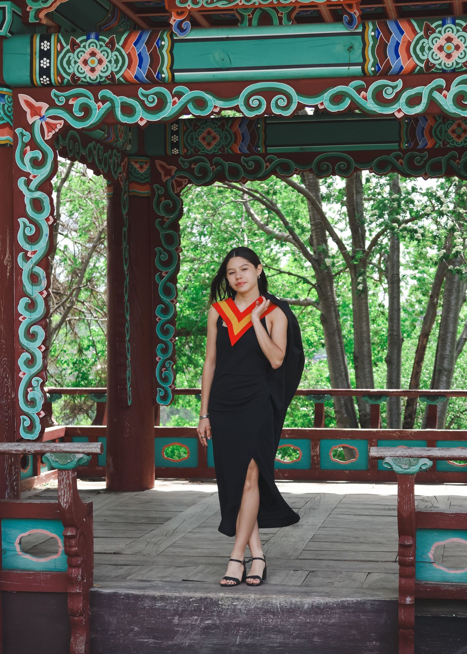 Young woman in black dress with graduation stole standing in colorful Asian pavilion.