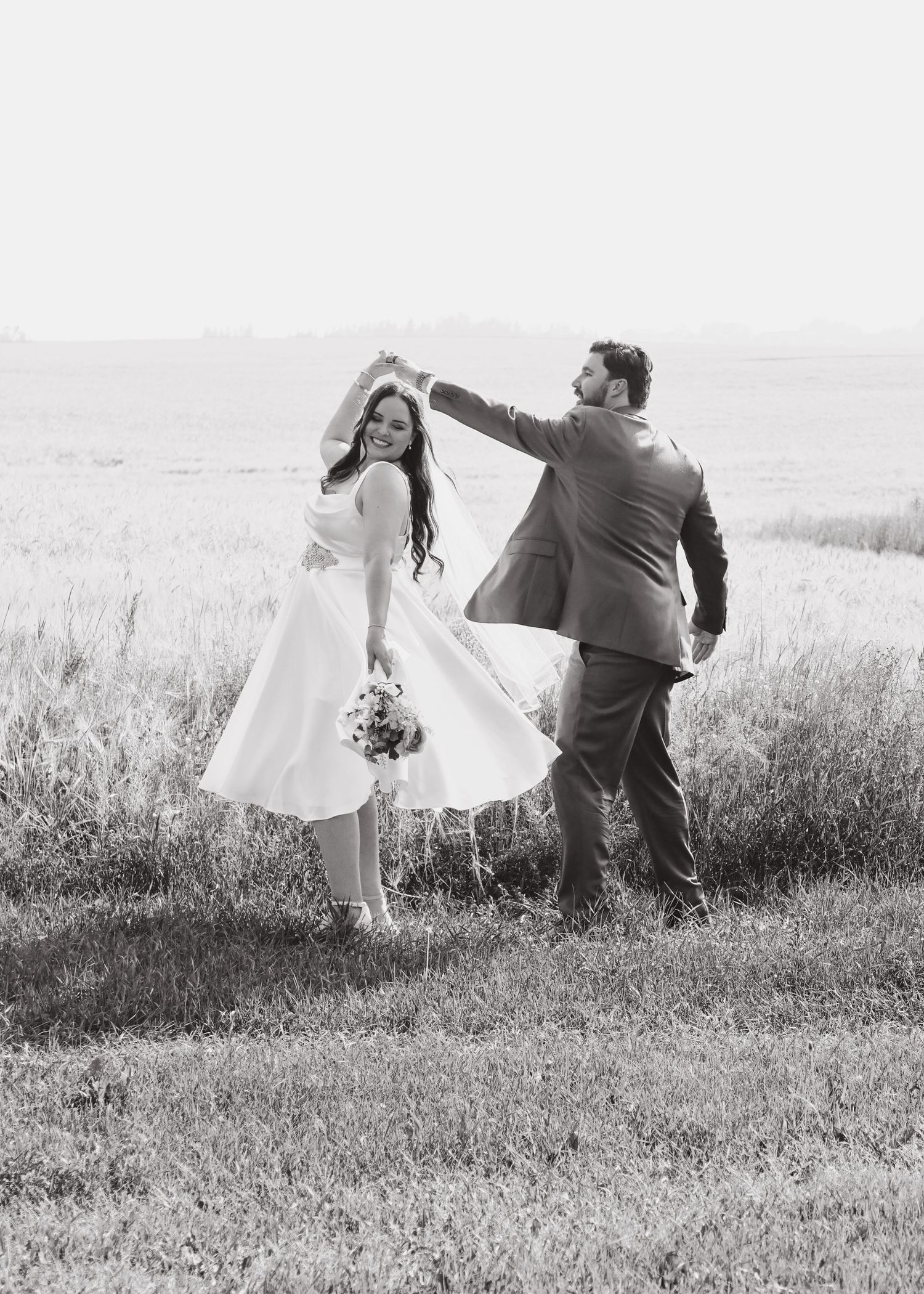 Black and white photo of groom spinning bride in white dress in open field.